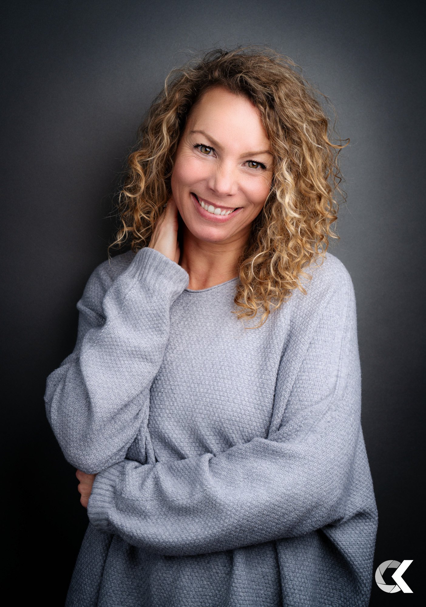 Portrait of a woman with curly blonde hair smiling, wearing a light gray sweatshirt, standing against a dark gray background.
