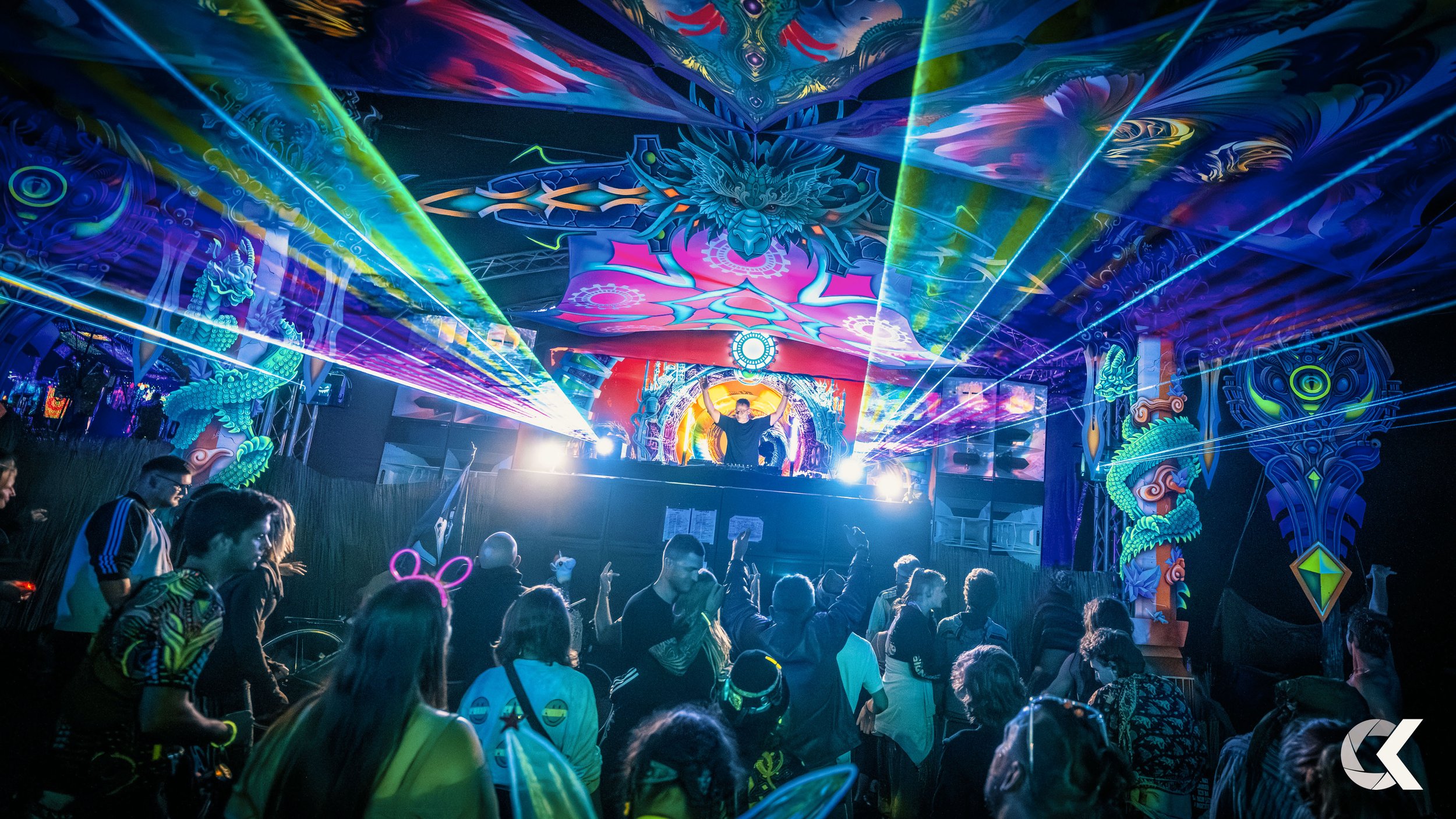 A DJ performing at a colorful electronic music event with neon lights, laser beams, and a crowd dancing.
