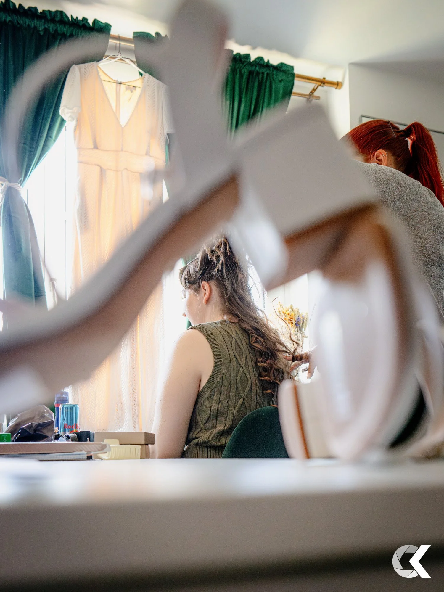 A woman with long, curly hair working with a makeup artist in a room with green curtains and a window. A white dress hangs in the background. The photo is taken from a low angle, through a pair of white high-heeled shoes.