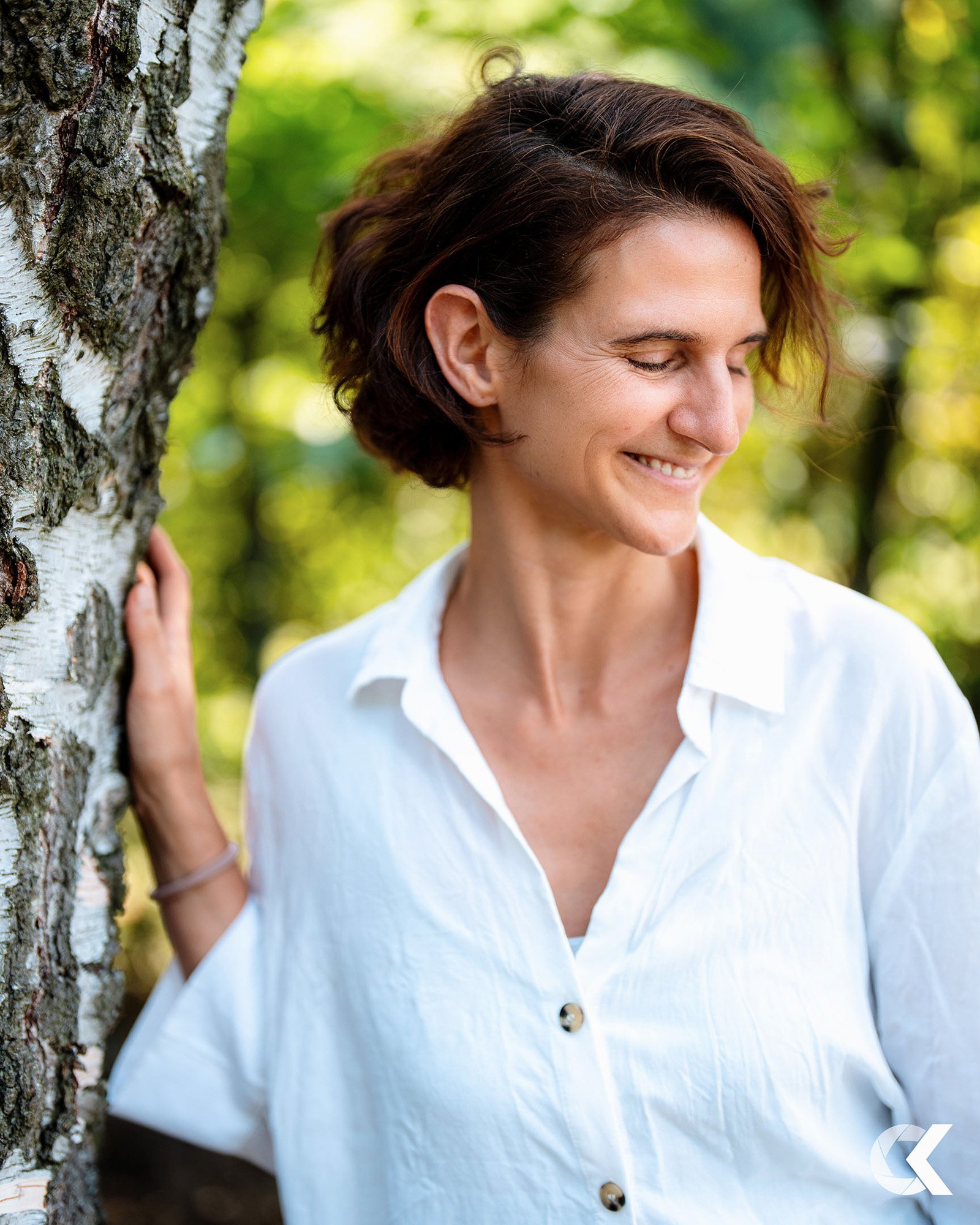 Freudestrahlende Frau lehnt an Baum im Wald bei sonnigem Wetter, trägt weiße Bluse.