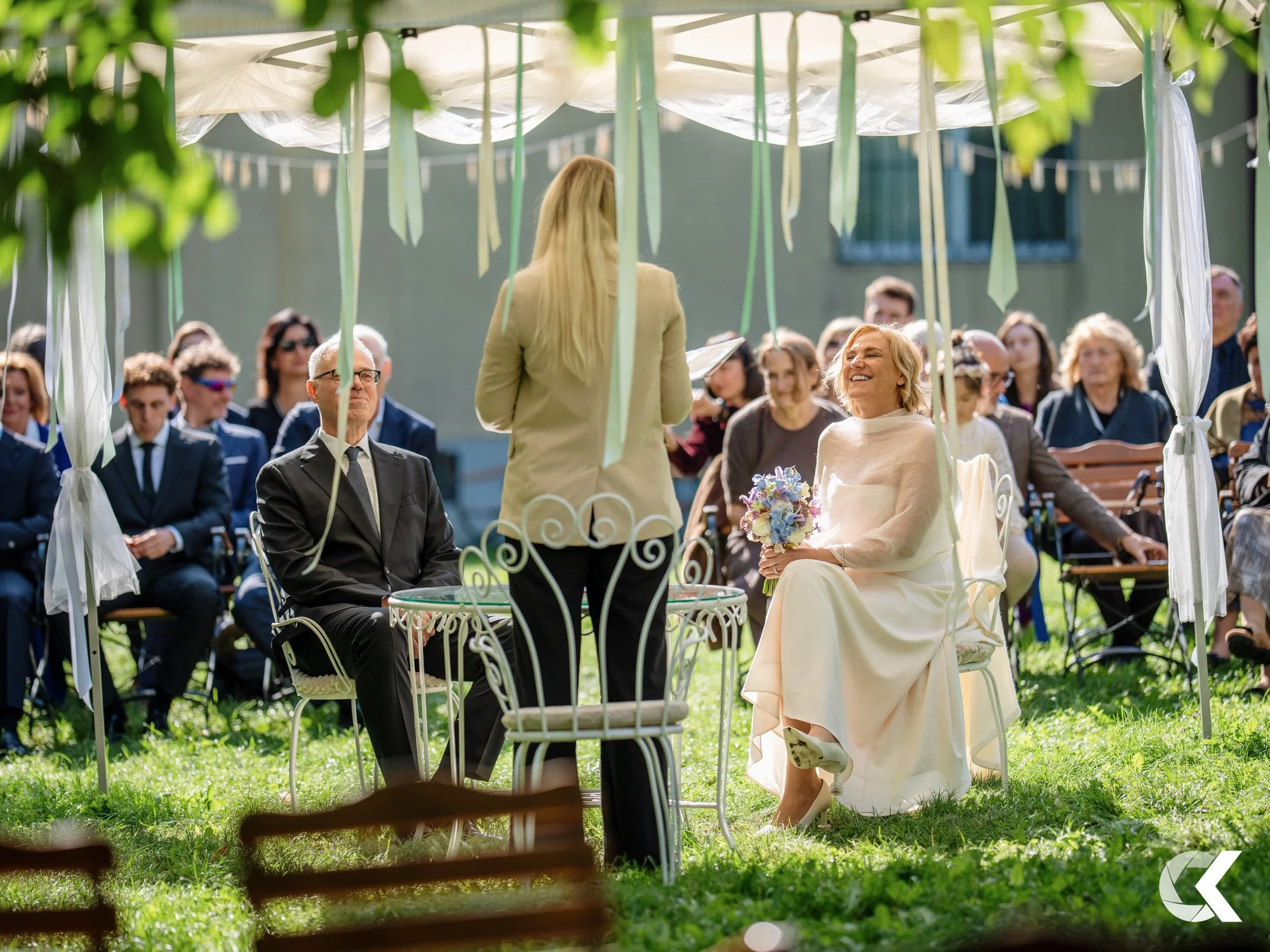 A wedding ceremony outdoors under a white canopy with ribbons. The bride in a white dress holds a bouquet and is seated, smiling at the officiant. An older man in a suit is seated next to her. Guests seated behind attend the ceremony on a sunny day.