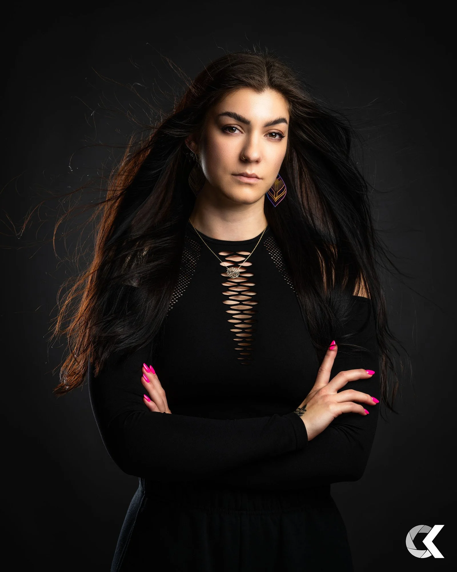 A young woman with long dark hair wearing a black long-sleeve top with cutout details in the front, standing against a dark background, with her arms crossed, pink painted nails, earrings, and a necklace.