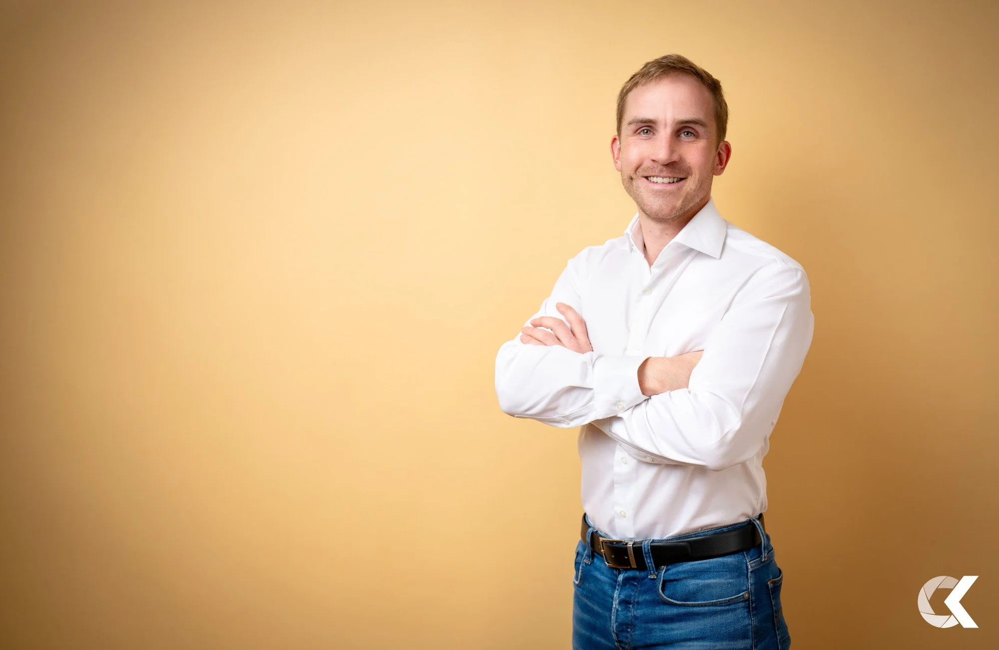 A smiling man with light brown hair, wearing a white dress shirt and blue jeans, standing with arms crossed against a yellow background.