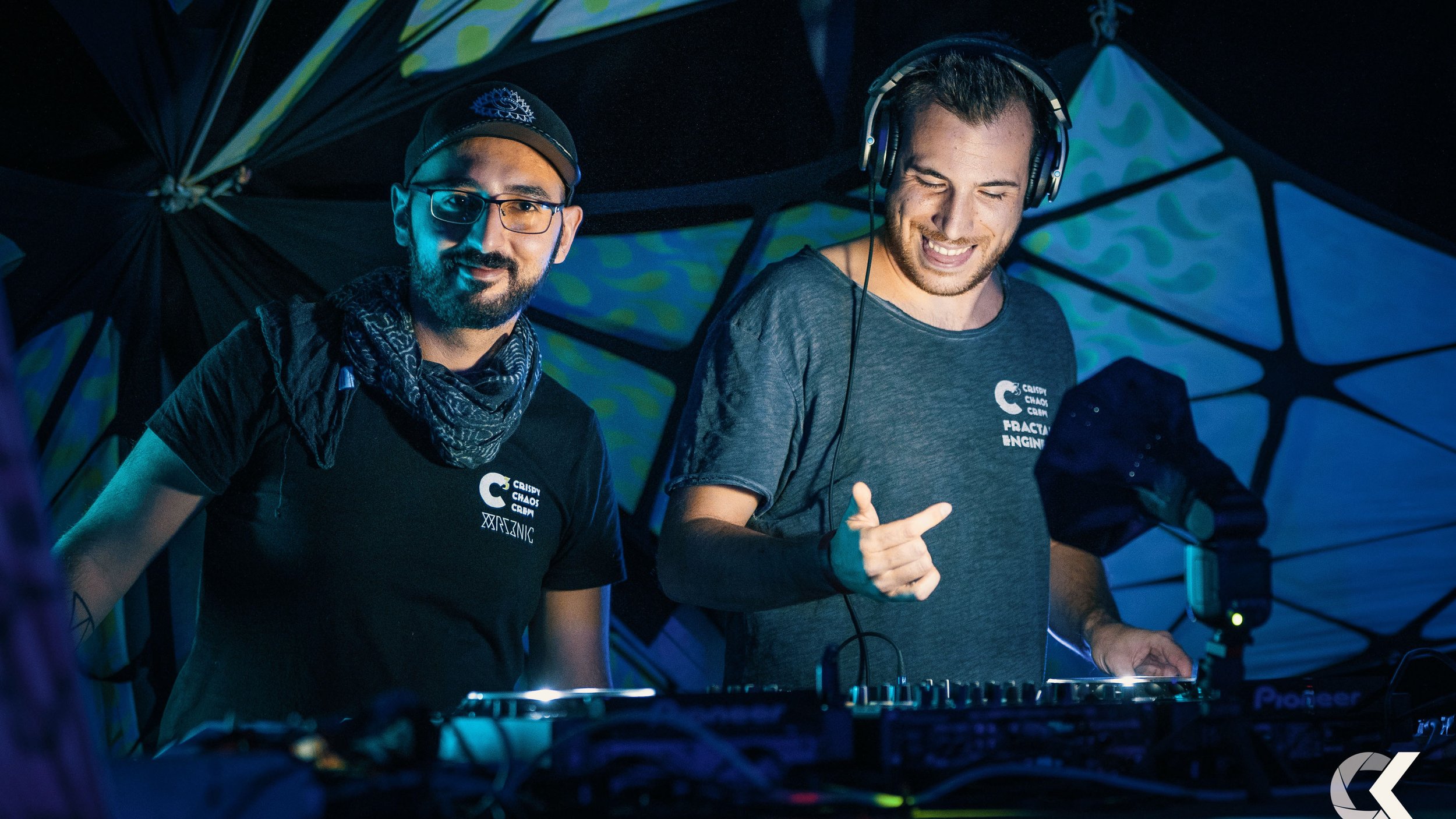 Two male DJs in a dark, colorful setting, one wearing glasses and a cap, the other with headphones, both smiling and standing behind DJ equipment.