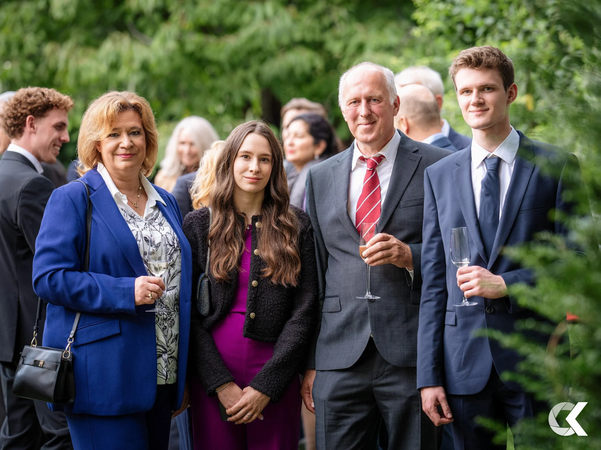 Group of four people, two women and two men, at outdoor social event, dressed in business attire, holding wine glasses, standing among other guests with greenery in background.
