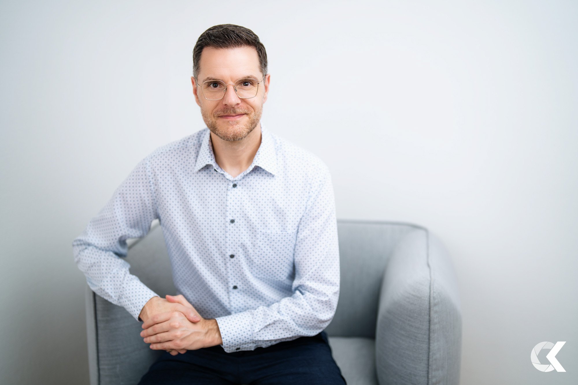 A man with glasses, light beard, and short brown hair sits on a light gray armchair. He is wearing a white button-up shirt with small dark polka dots and dark pants, looking directly at the camera with a slight smile, in front of a plain white backgr