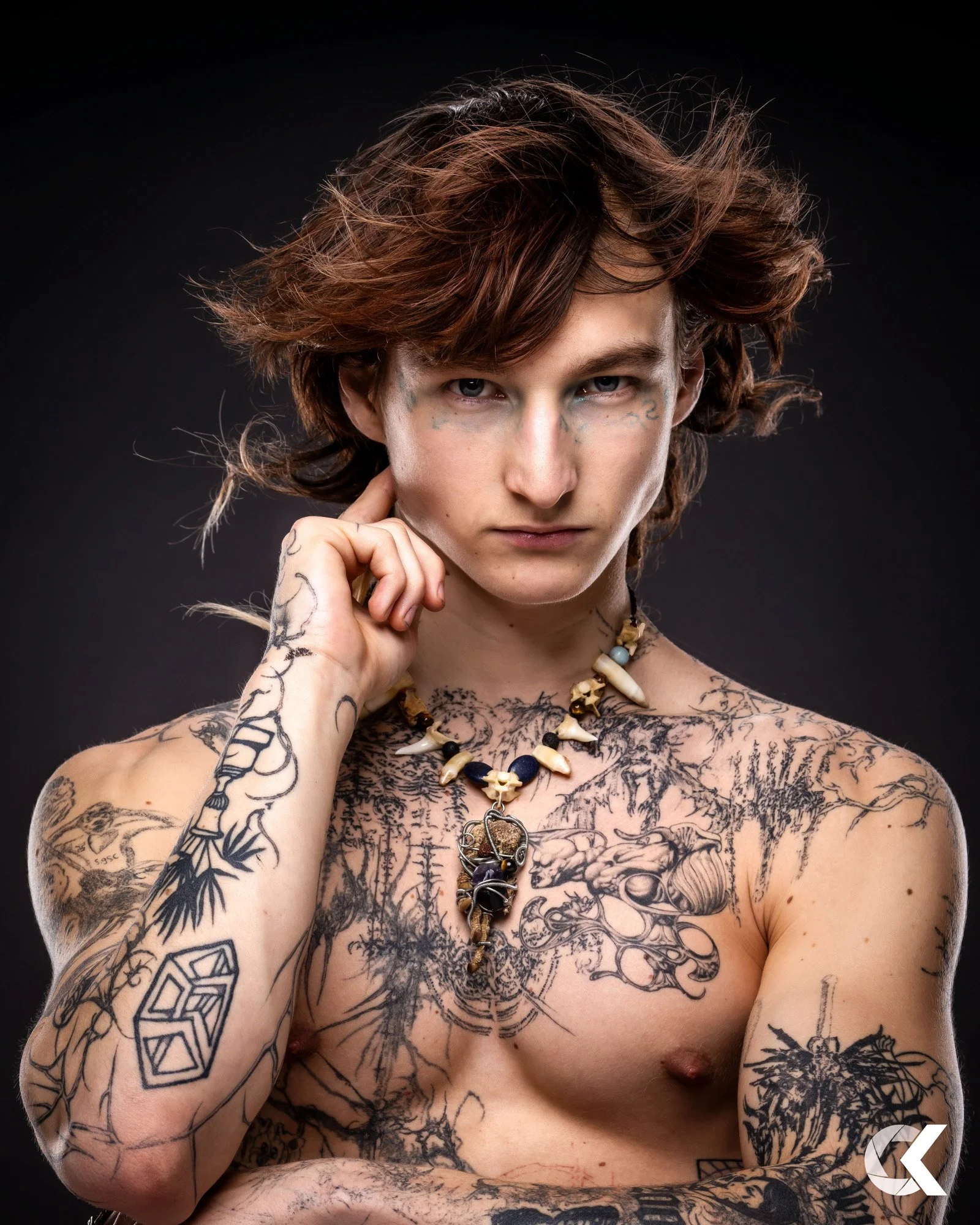 A young man with shoulder-length wavy brown hair and tattoos on his arms and chest, wearing a necklace with beads and a pendant, looking directly at the camera against a dark background.