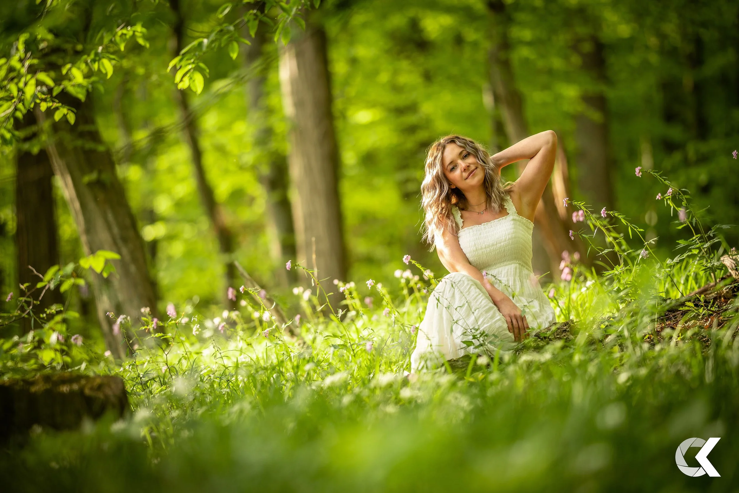 A young woman in a white dress sitting on the grass in a lush green forest with sunlight filtering through the trees.