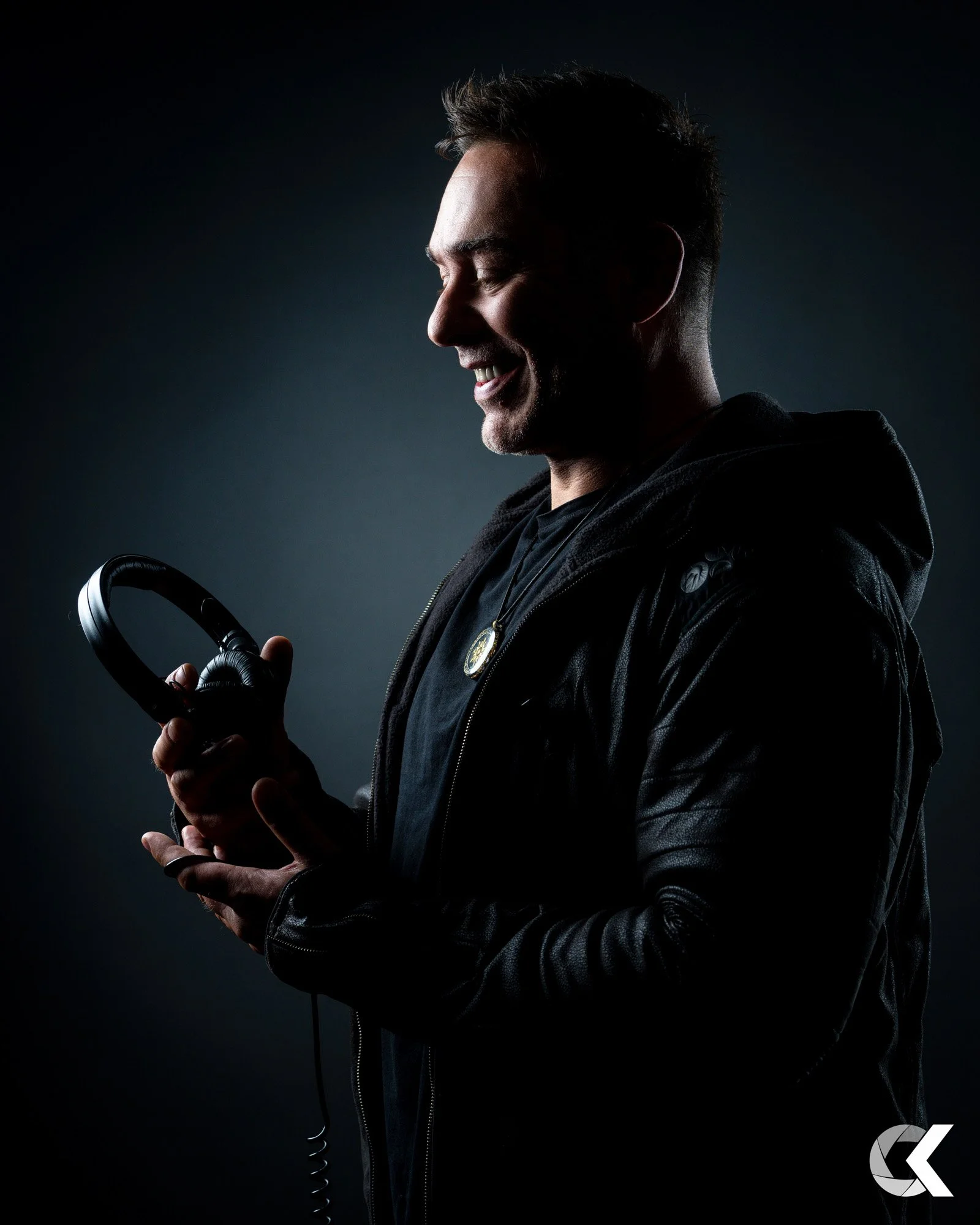 Ein lachender Mann trägt eine schwarze Jacke und hält Kopfhörer in der Hand, vor einem dunklen Hintergrund.