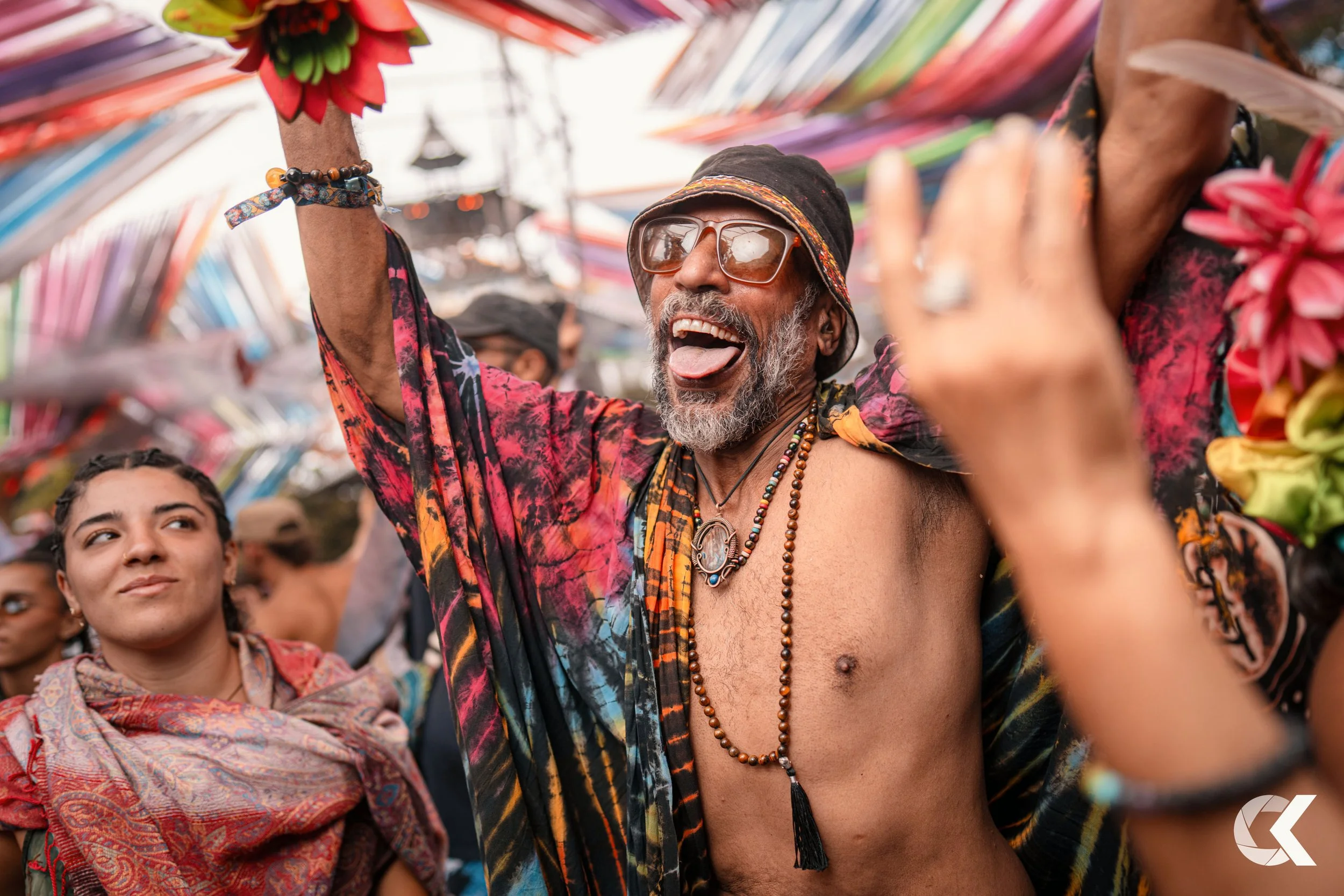 A joyful man with gray hair, beard, sunglasses, and colorful clothing, dancing among a crowd at a festival with vibrant decorations overhead.