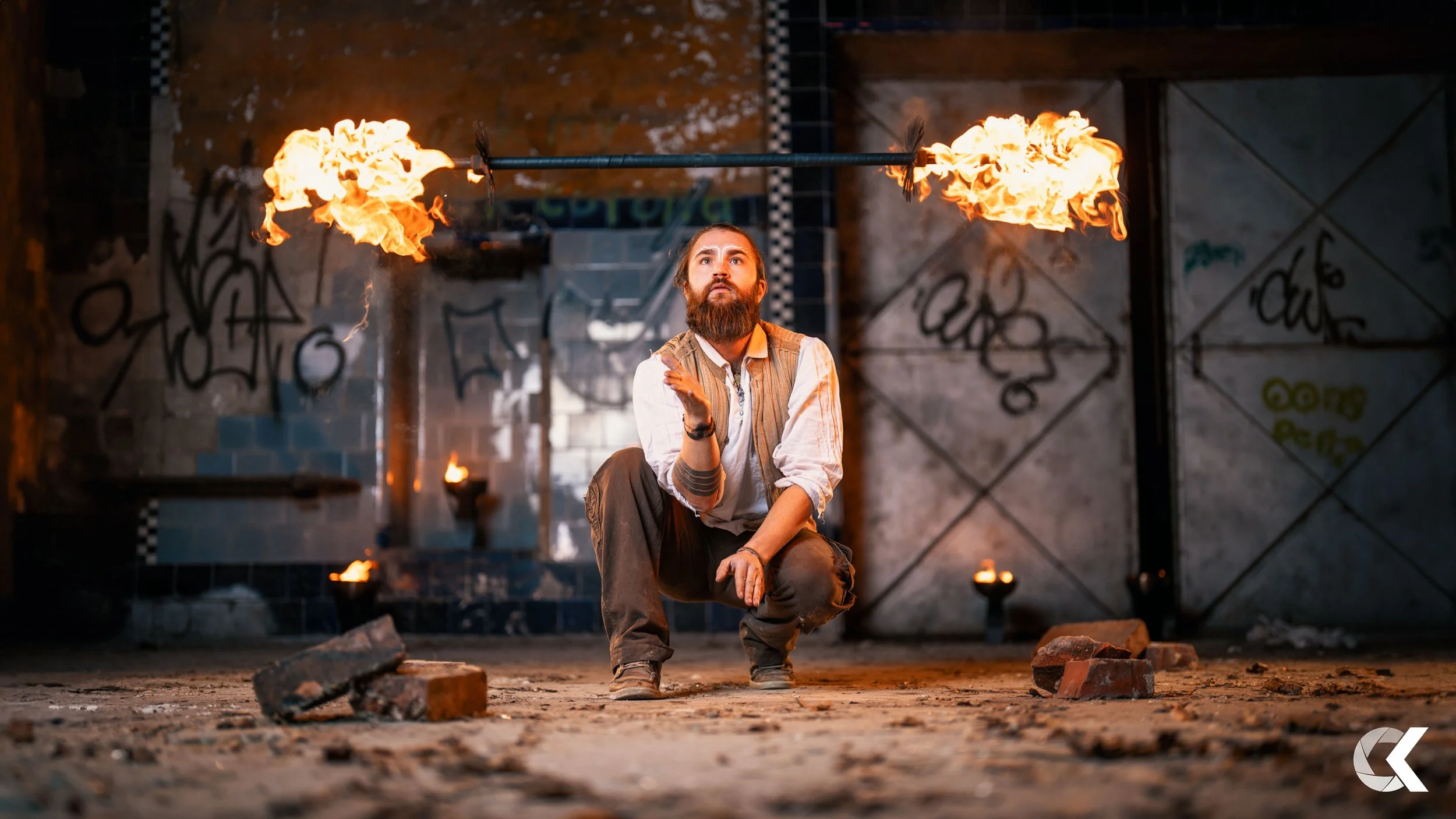 A man with a beard and long hair, dressed in a light shirt and dark pants, is kneeling on the floor of an industrial-style space as he spins a fire staff horizontally above his head, creating flames on both ends. The background includes graffiti on t