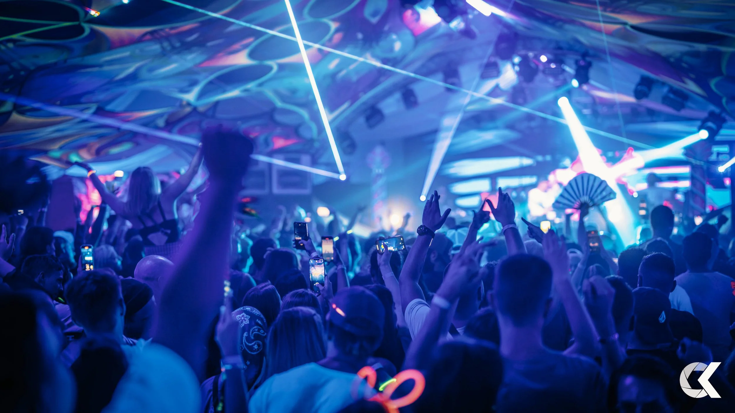 A lively concert scene with a large crowd of people dancing and taking photos, illuminated by colorful stage lights and laser beams.