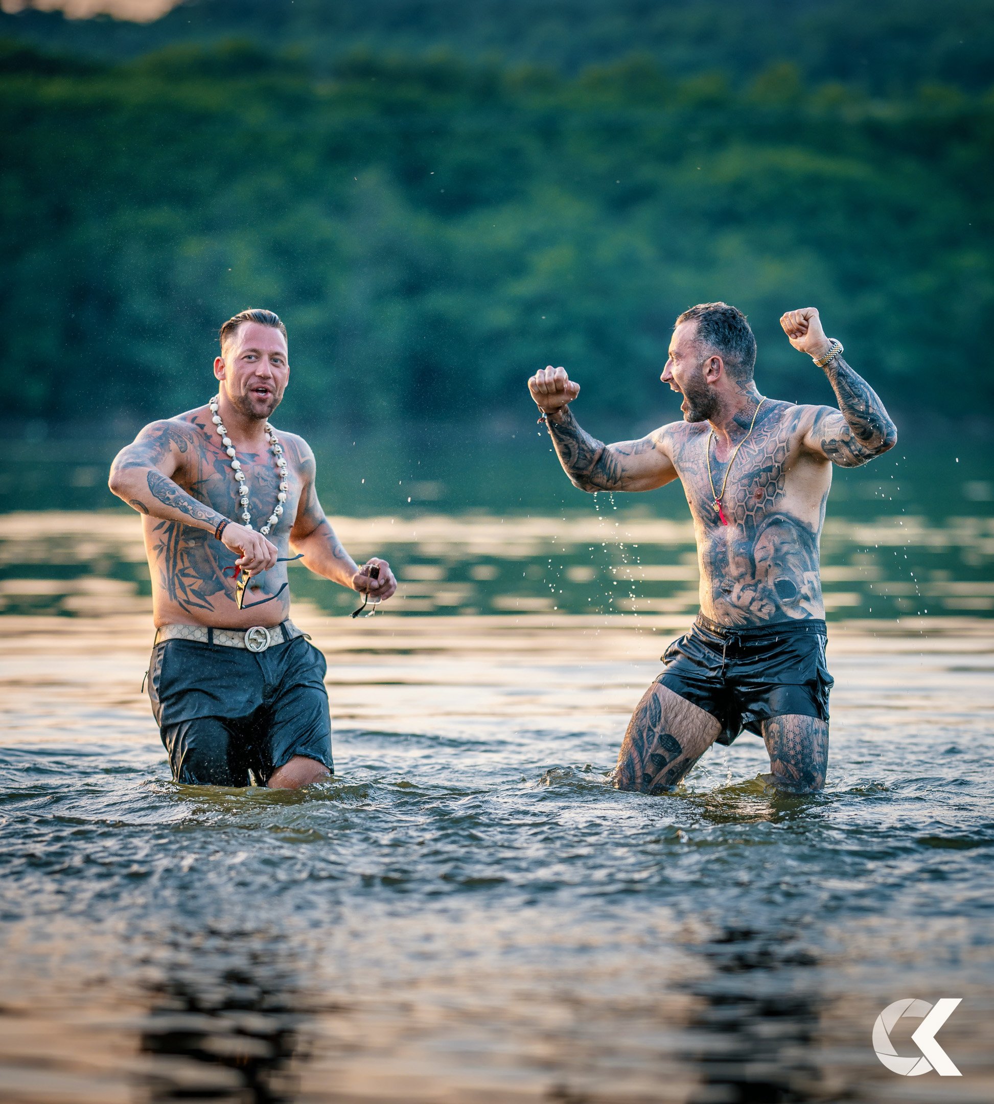 Two tattooed men in black swim trunks enjoying a swim in a lake, with one smiling and the other flexing his muscles, all set against a lush green forest background during sunset.