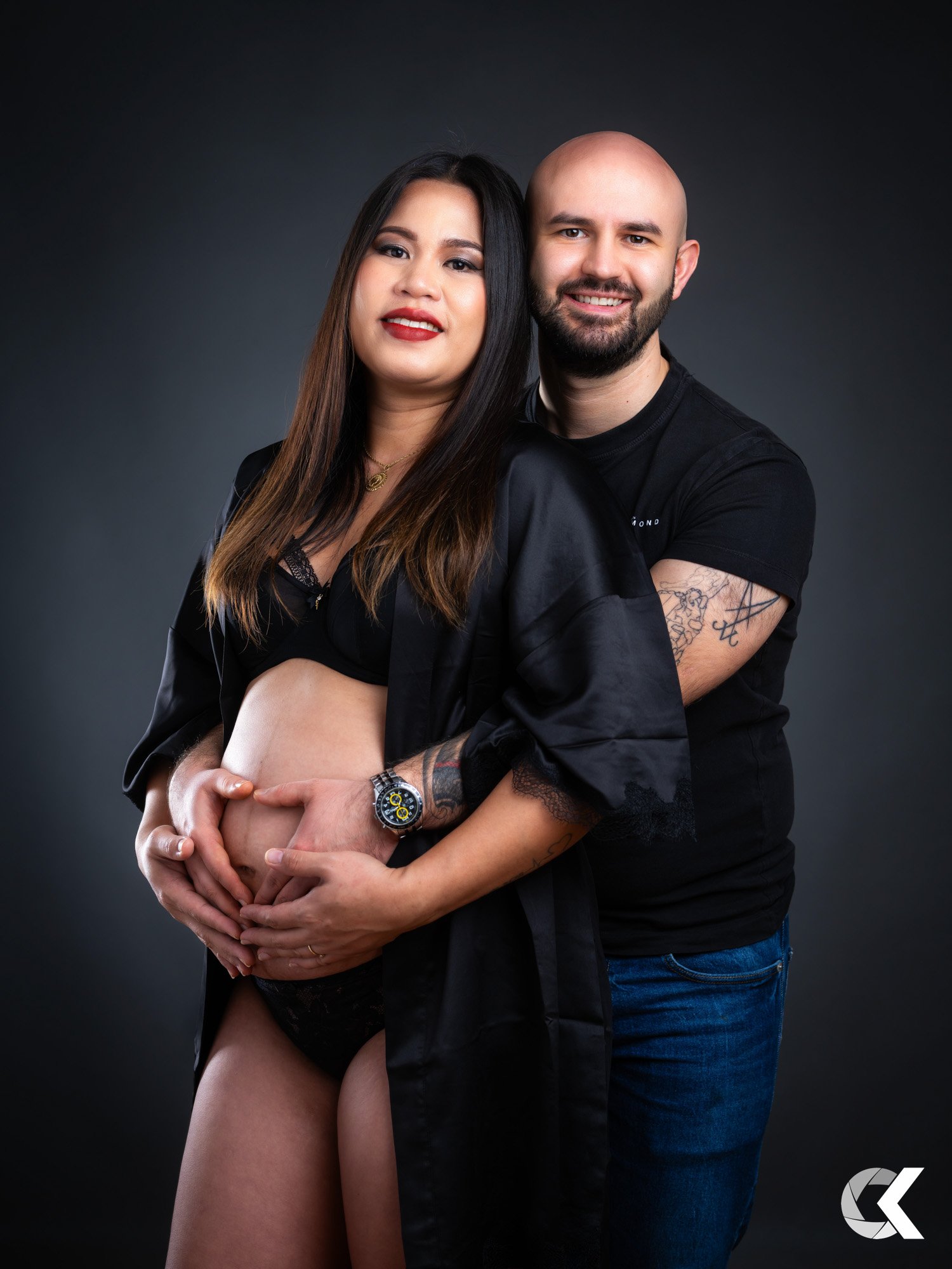 A pregnant woman and her partner stand closely together, with their hands forming a heart shape over her baby bump, against a black background.