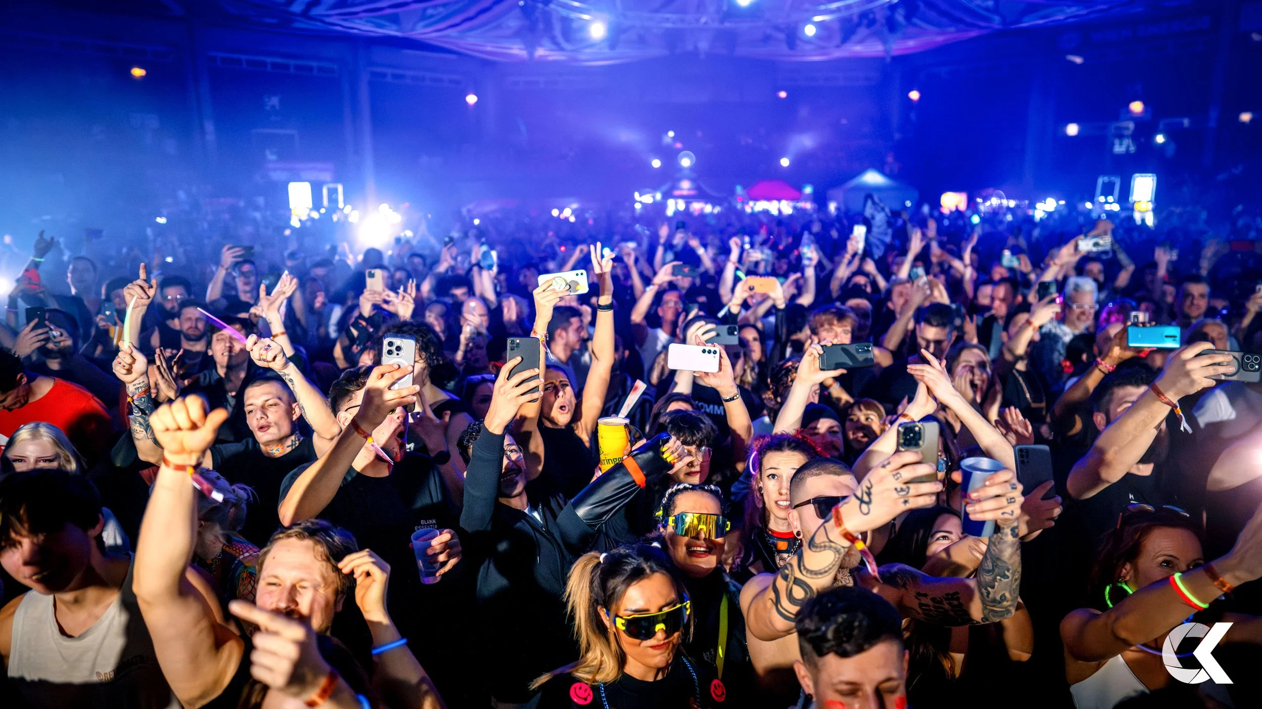 Crowd at a concert or music event, many people holding up smartphones to record or take pictures, colorful lighting and a lively atmosphere