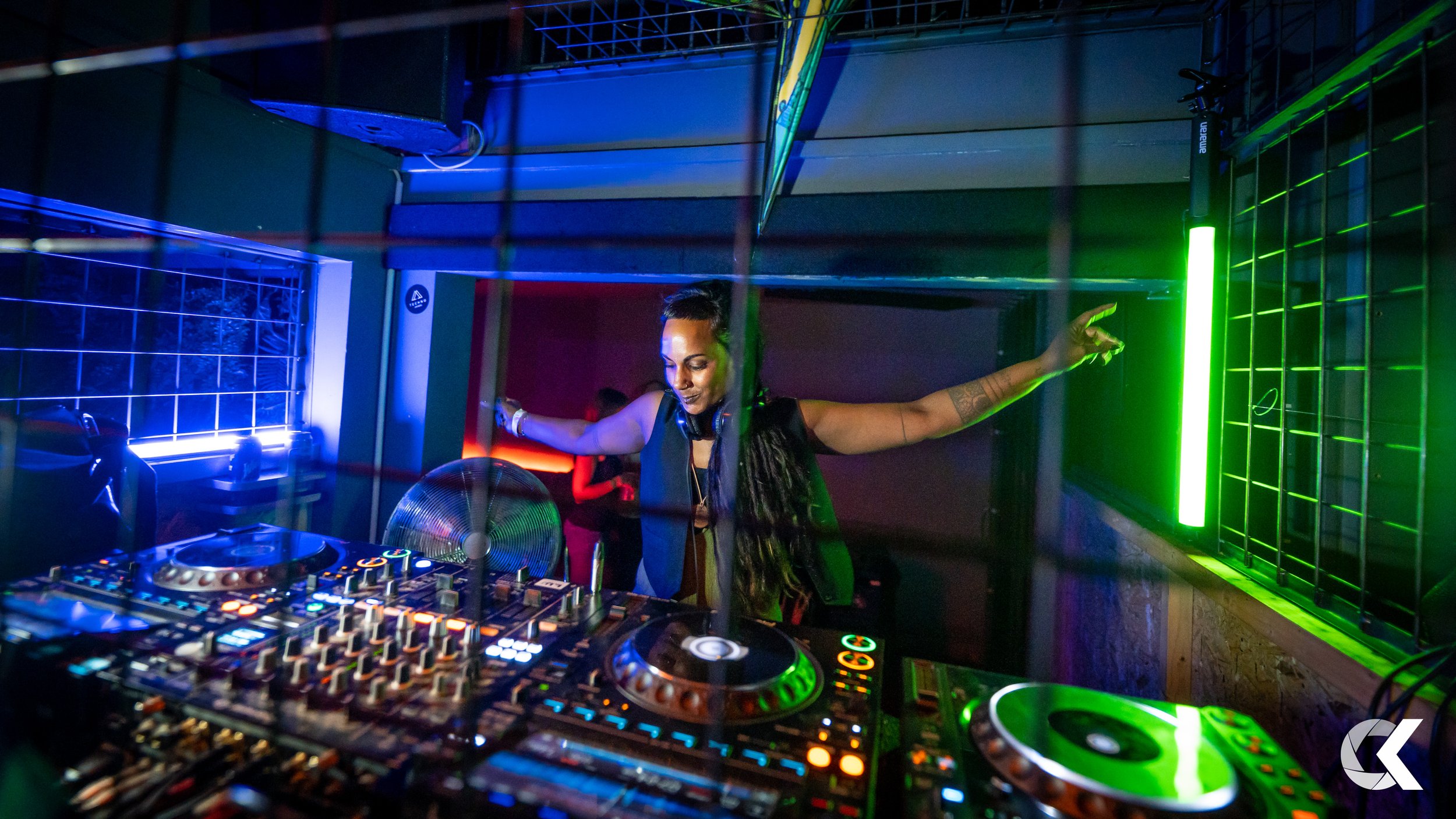 A female DJ with long braids, wearing headphones around her neck, standing behind a DJ setup with turntables and mixers, in a dimly lit nightclub with green and blue lighting.