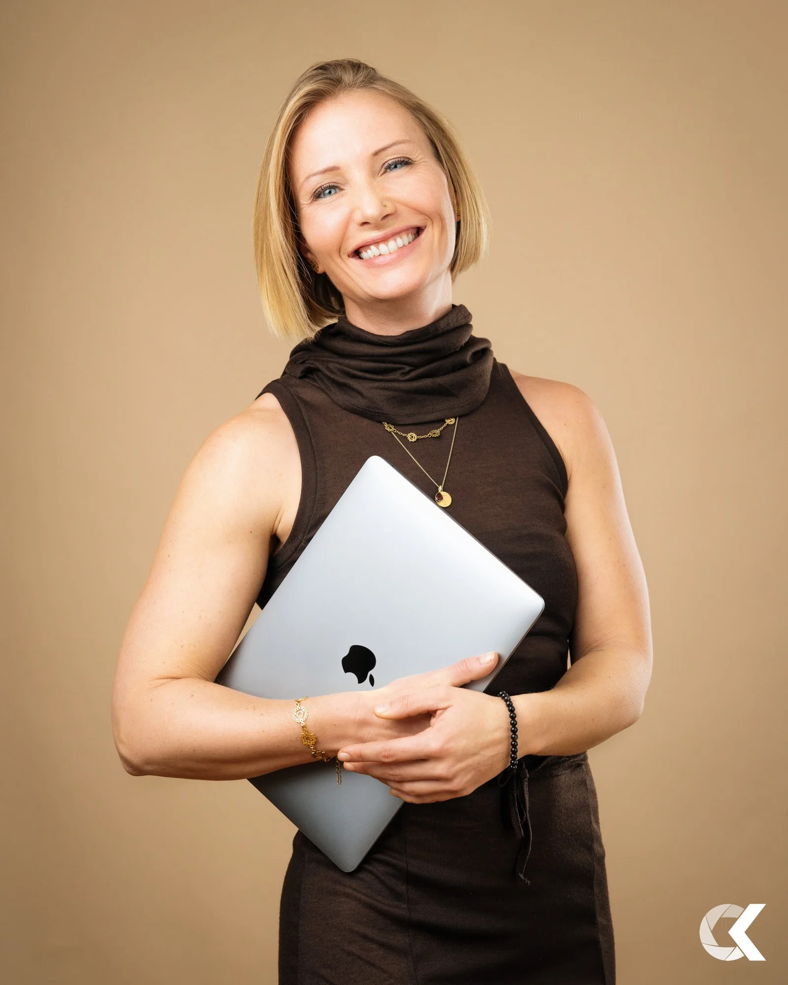 Smiling woman with short blonde hair holding a closed Apple MacBook, wearing a sleeveless black top, gold jewelry, and a bracelet, against a beige background.