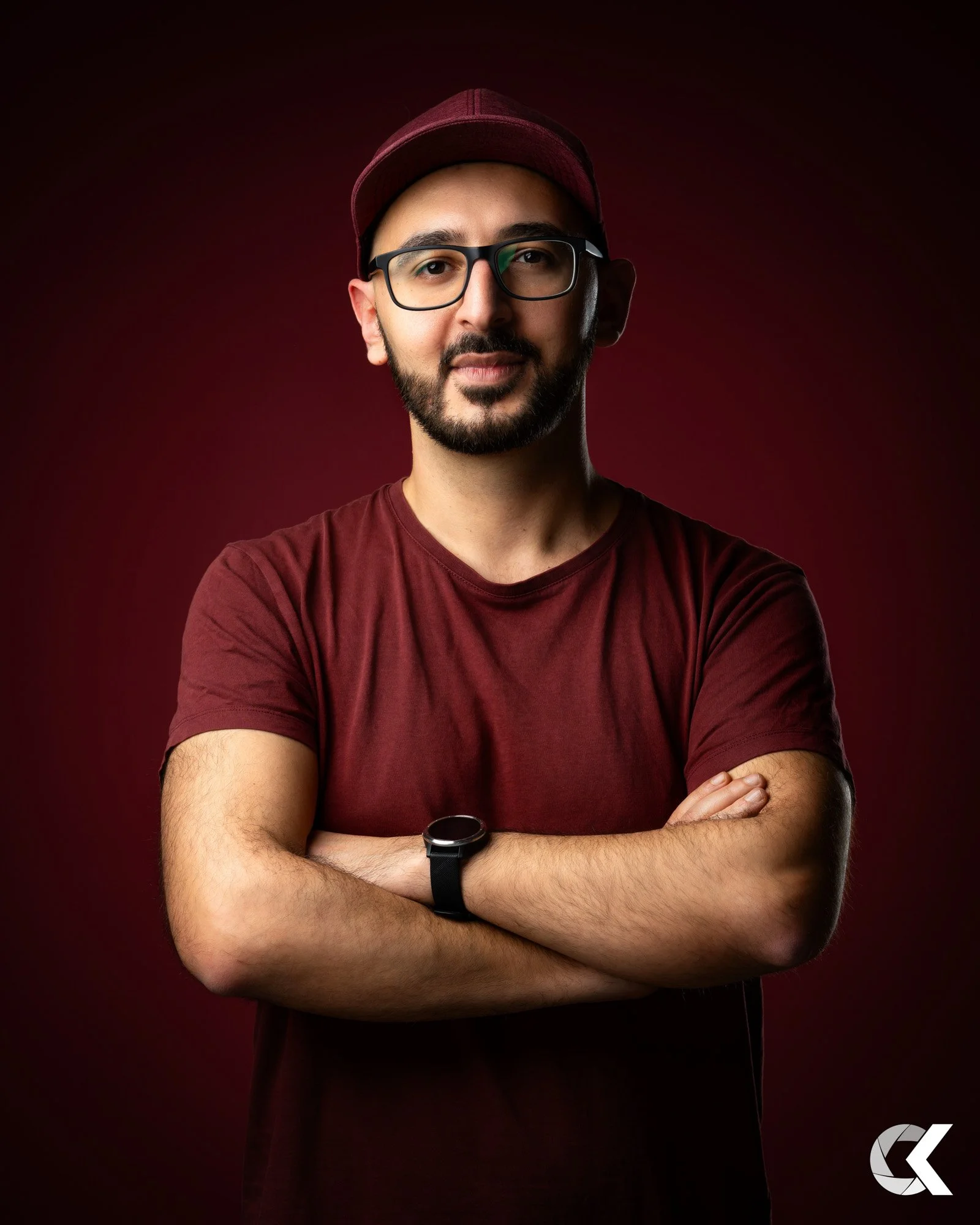 Ein Mann mit dunklem Bart, Brille und roten Kleidung steht vor dunklem roten Hintergrund, Arme verschränkt, trägt eine Armbanduhr, schaut in die Kamera.