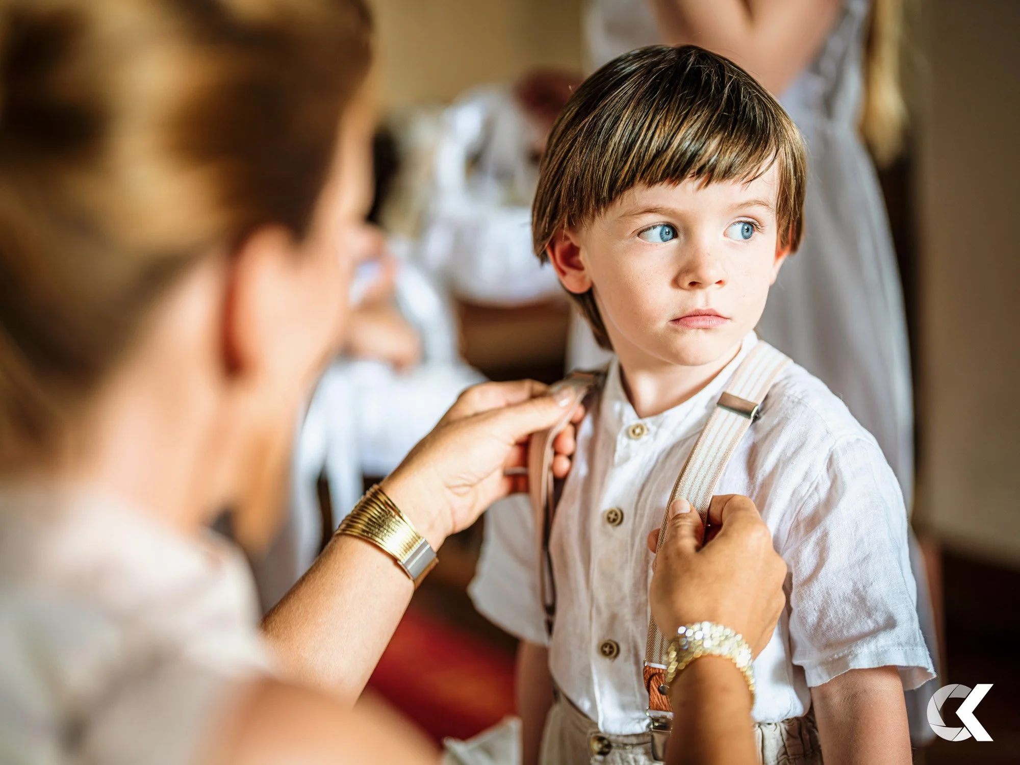 An elderly woman helps a young boy put on suspenders in a warm, indoor setting.