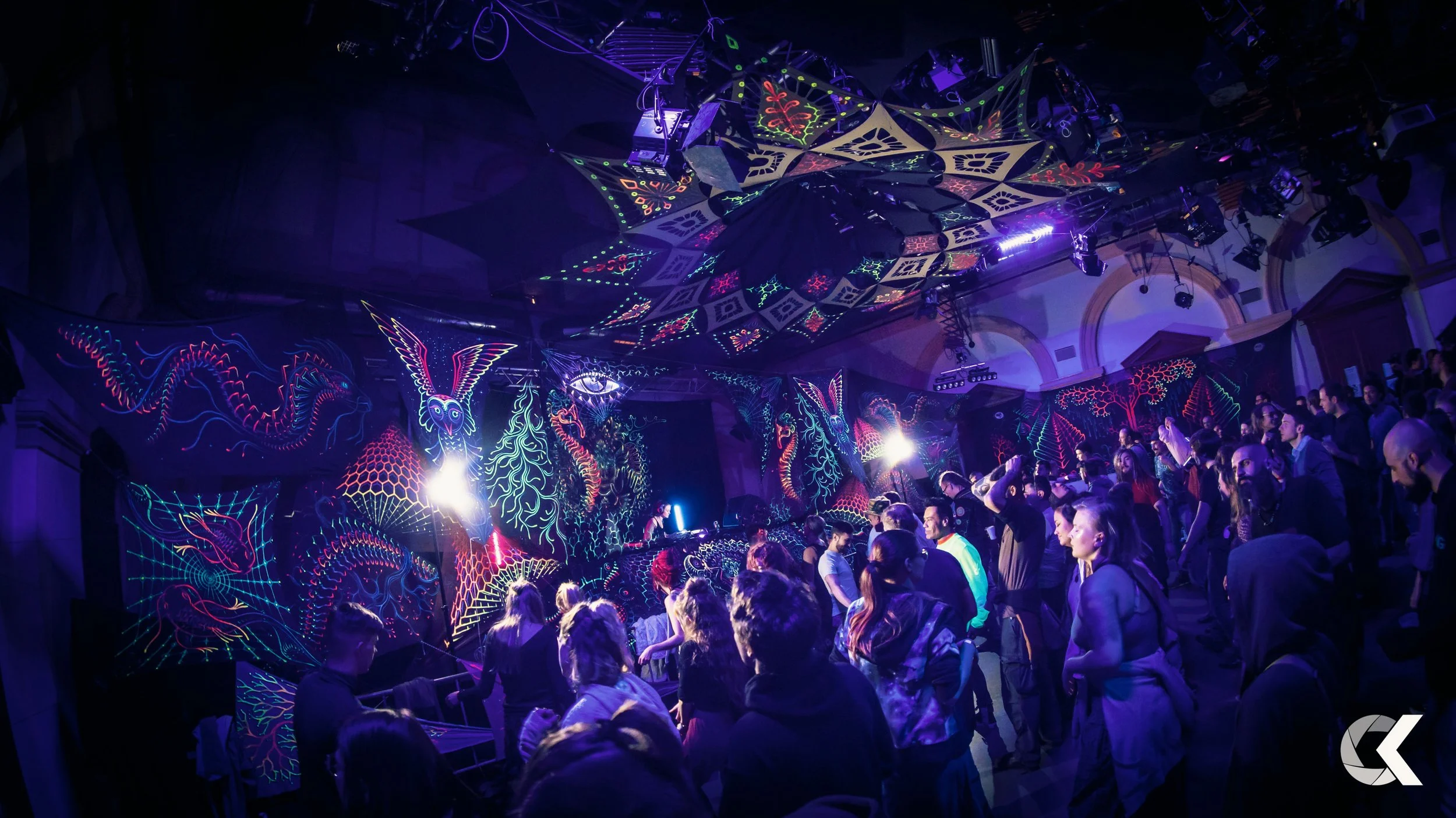 A crowded indoor party with neon, glow-in-the-dark, or blacklight-illuminated artwork on the walls and ceiling, featuring abstract and fantasy designs, with people dancing and socializing.