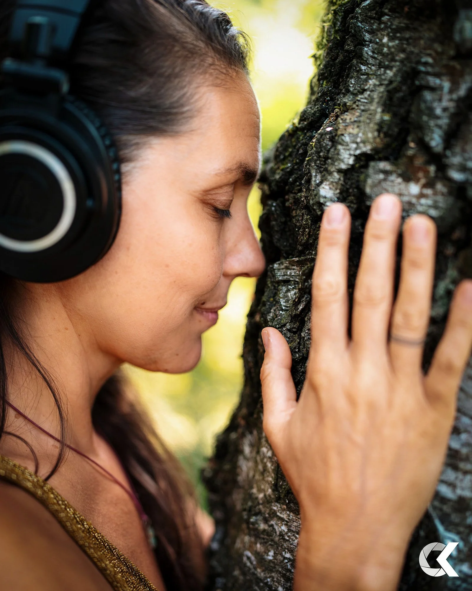 A woman with closed eyes, wearing headphones, touching a tree trunk with her hand, in a forest setting.