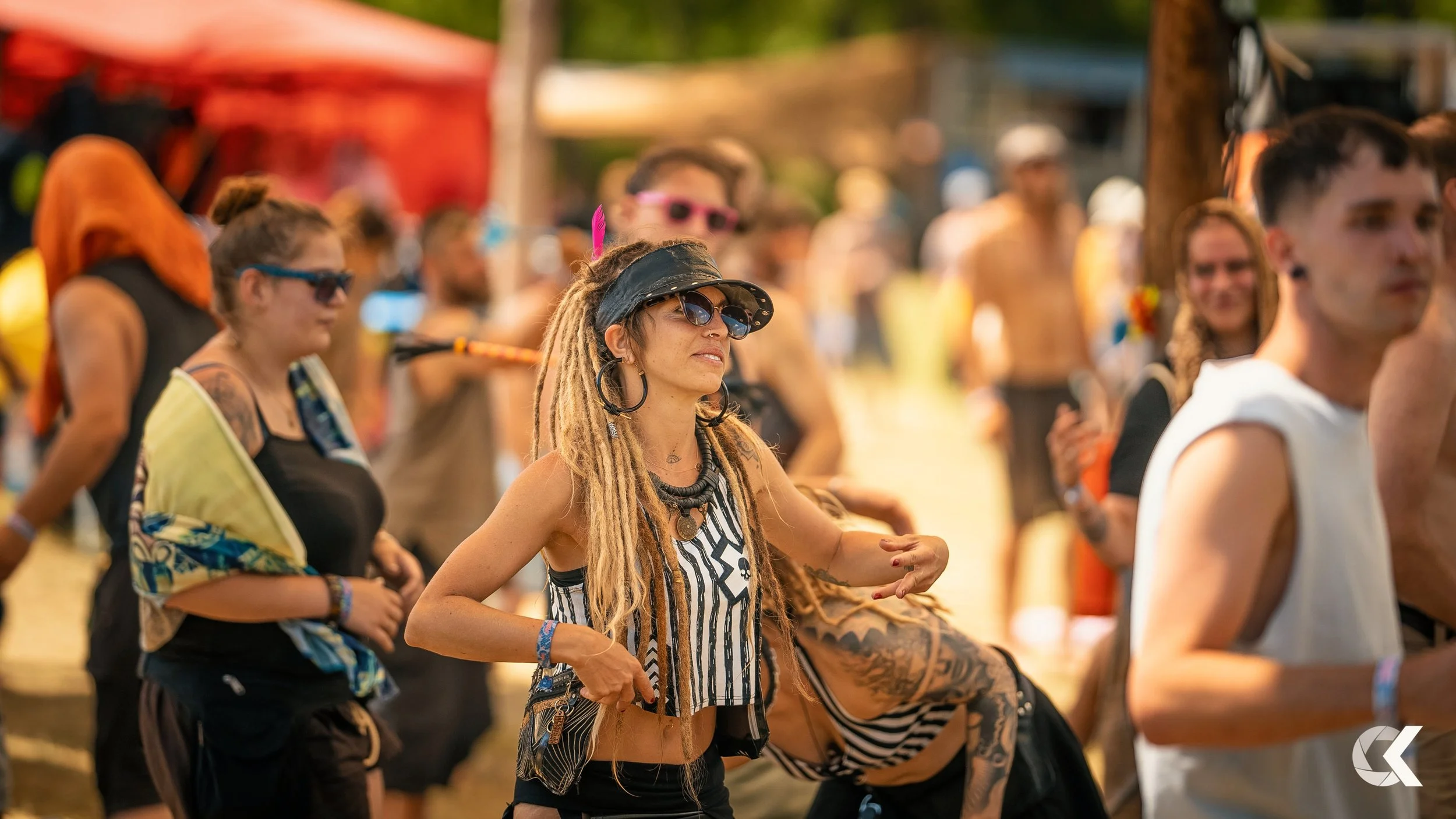 A group of people at an outdoor music festival, with a woman with dreadlocks, sunglasses, and a hat in focus, wearing a striped crop top and jewelry, among other festival-goers in casual attire.