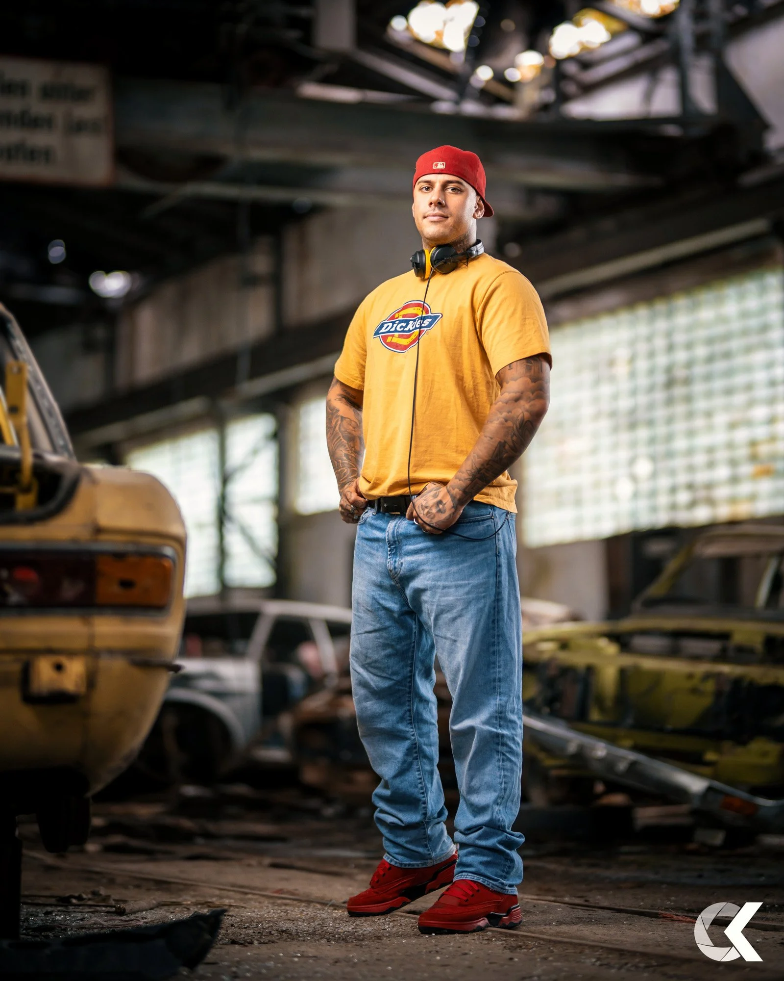 Junger Mann steht in einer alten, verfallenen Autofabrik, trägt ein gelbes T-Shirt, blaue Jeans, rote Schuhe und eine rote Mütze. Er hat Tätowierungen an den Armen.