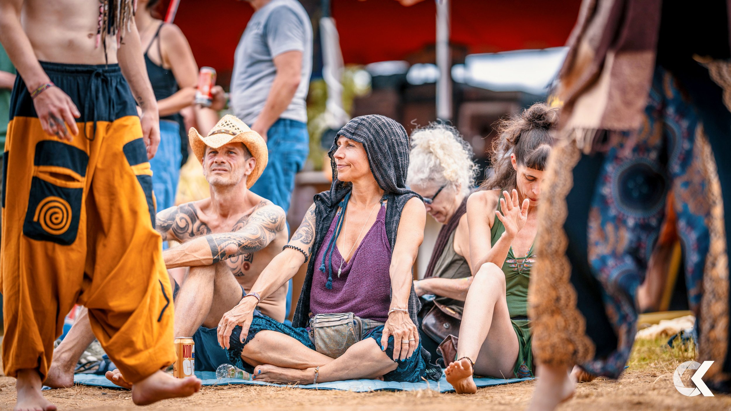 People sitting and standing outdoors during a gathering or festival with a red tent in the background. Some have tattoos, tattoos, and a woman with curly white hair and glasses, others are dressed casually, with some in swimwear or bohemian clothing.