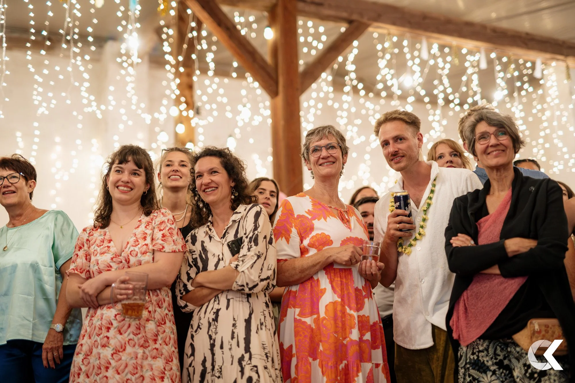 Group of smiling people at a party or celebration under hanging string lights, some holding drinks, in a decorated indoor venue.