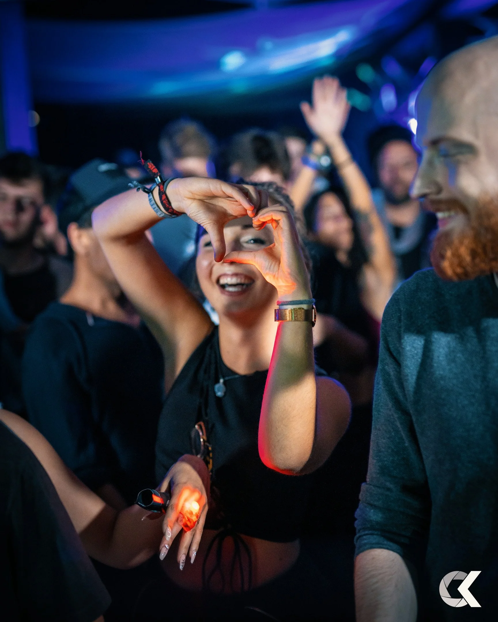 A group of people dancing at a party, with a smiling woman making a heart shape with her hands in the foreground.