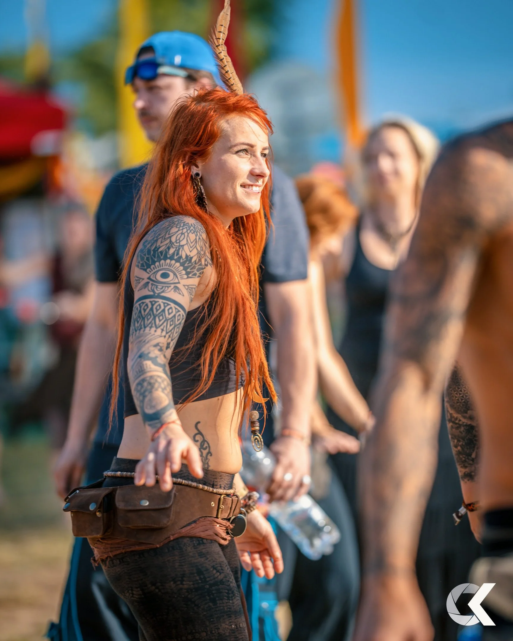A woman with long red hair and numerous tattoos on her arms, wearing a crop top and lace pants, smiling and engaging with others at an outdoor event.