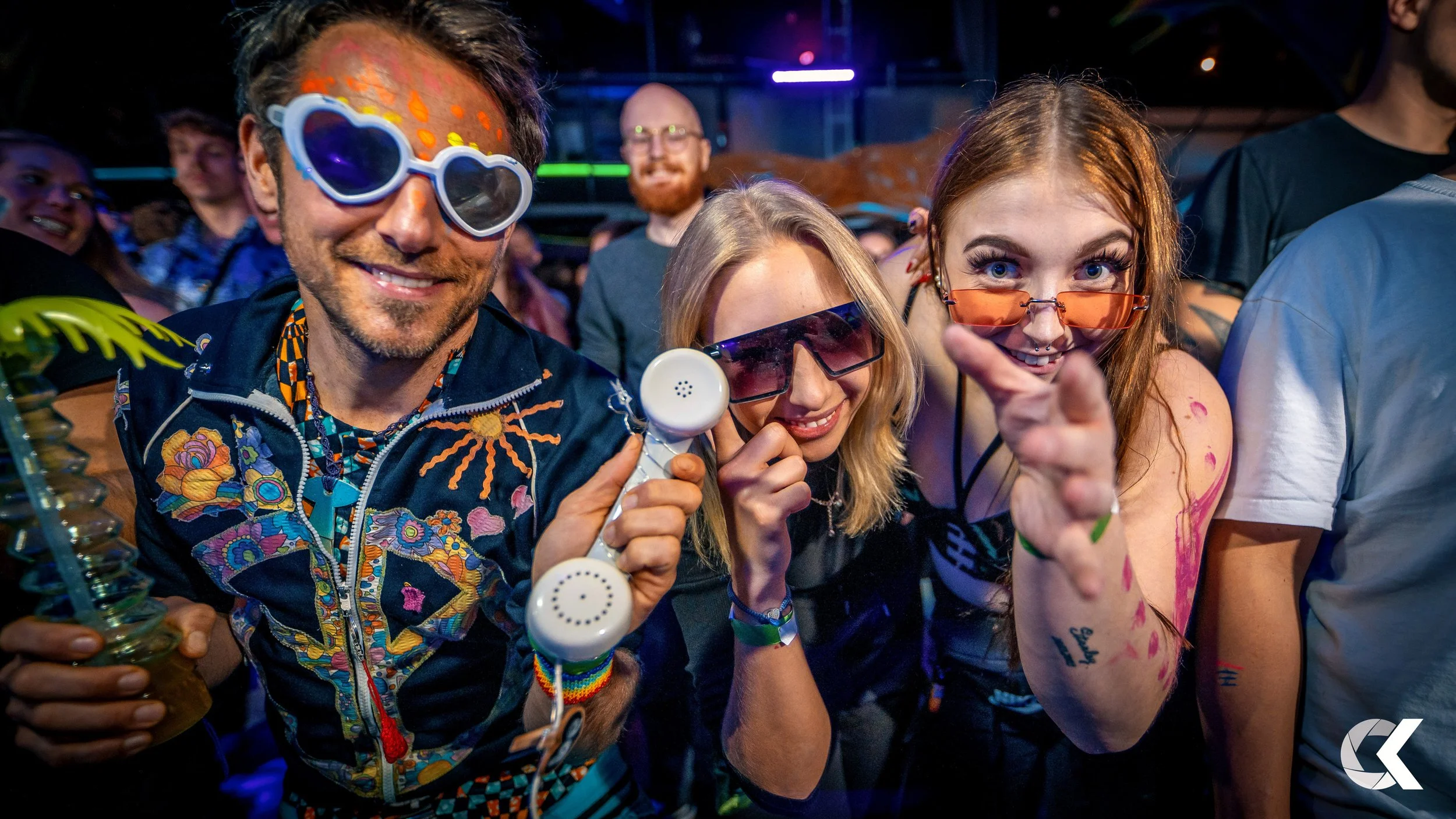 Group of young adults at a lively party wearing colorful sunglasses, with a man holding a vintage telephone, smiling and posing for the camera.