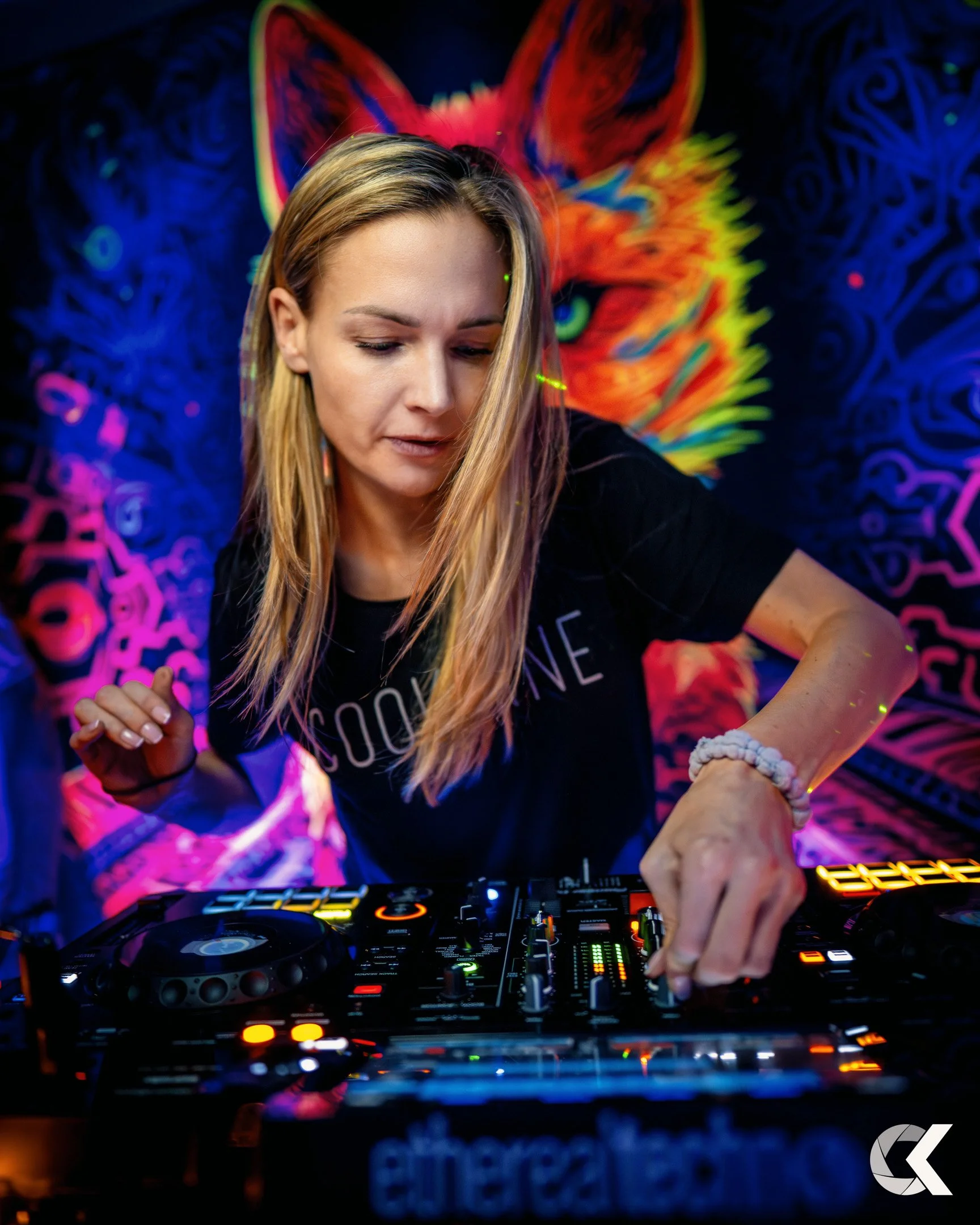 A blonde female DJ wearing a black t-shirt, mixing music on a DJ controller with colorful LED lights, with a vibrant, neon-colored wolf's face artwork in the background.