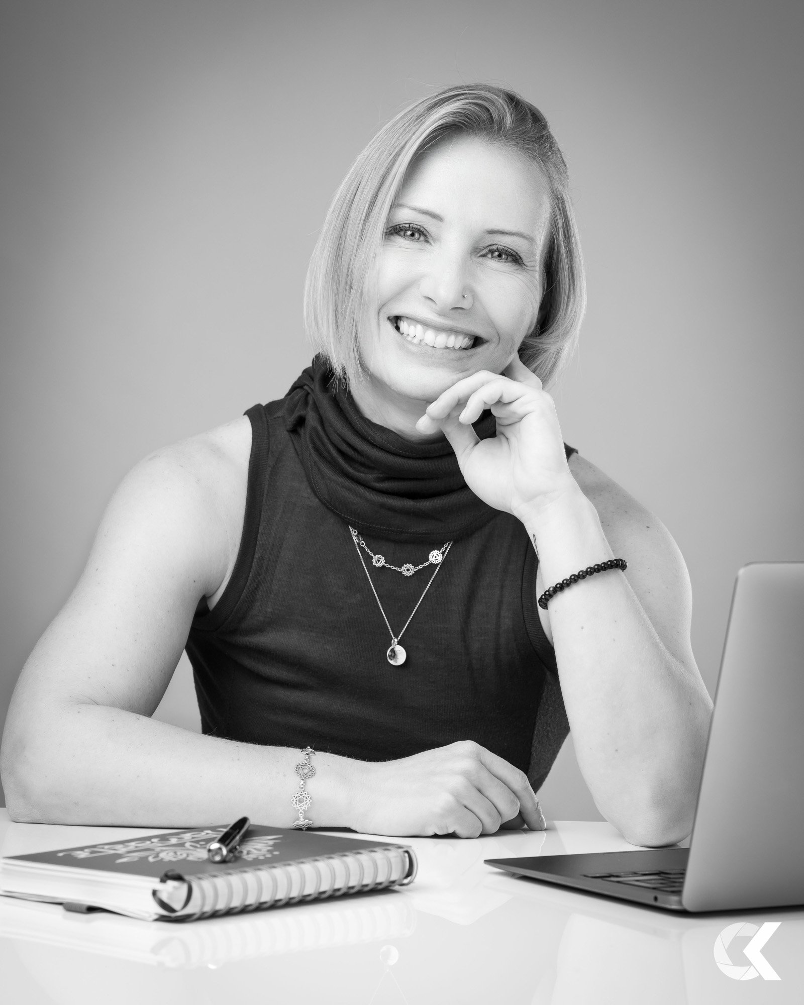 Black and white photo of a smiling woman with short hair, wearing a sleeveless top, jewelry, and a necklace, sitting at a desk with a laptop and notebooks.