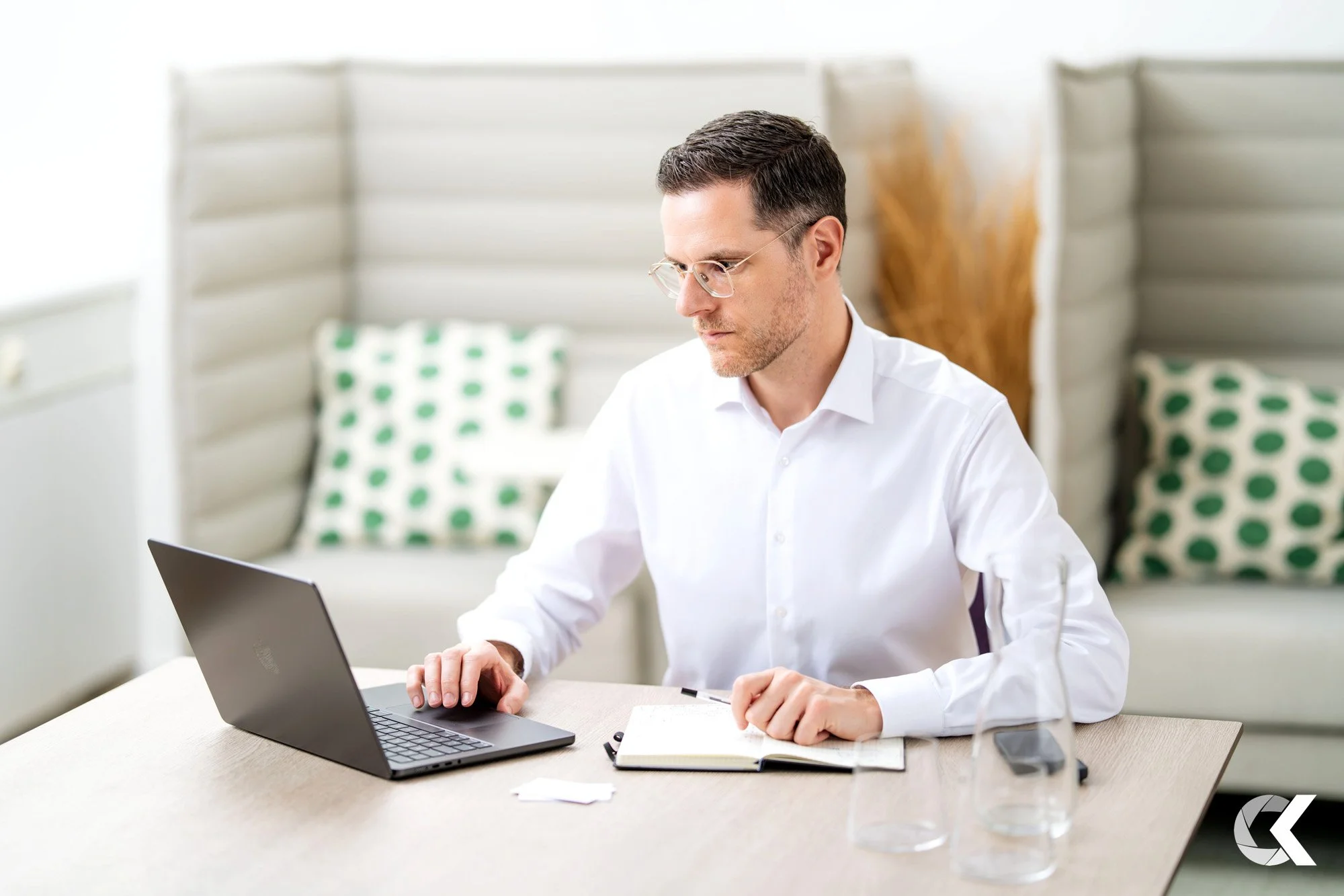 A man with glasses and a white shirt working on a laptop at a desk with a notebook, a pen, and two glasses of water, in a modern office setting.