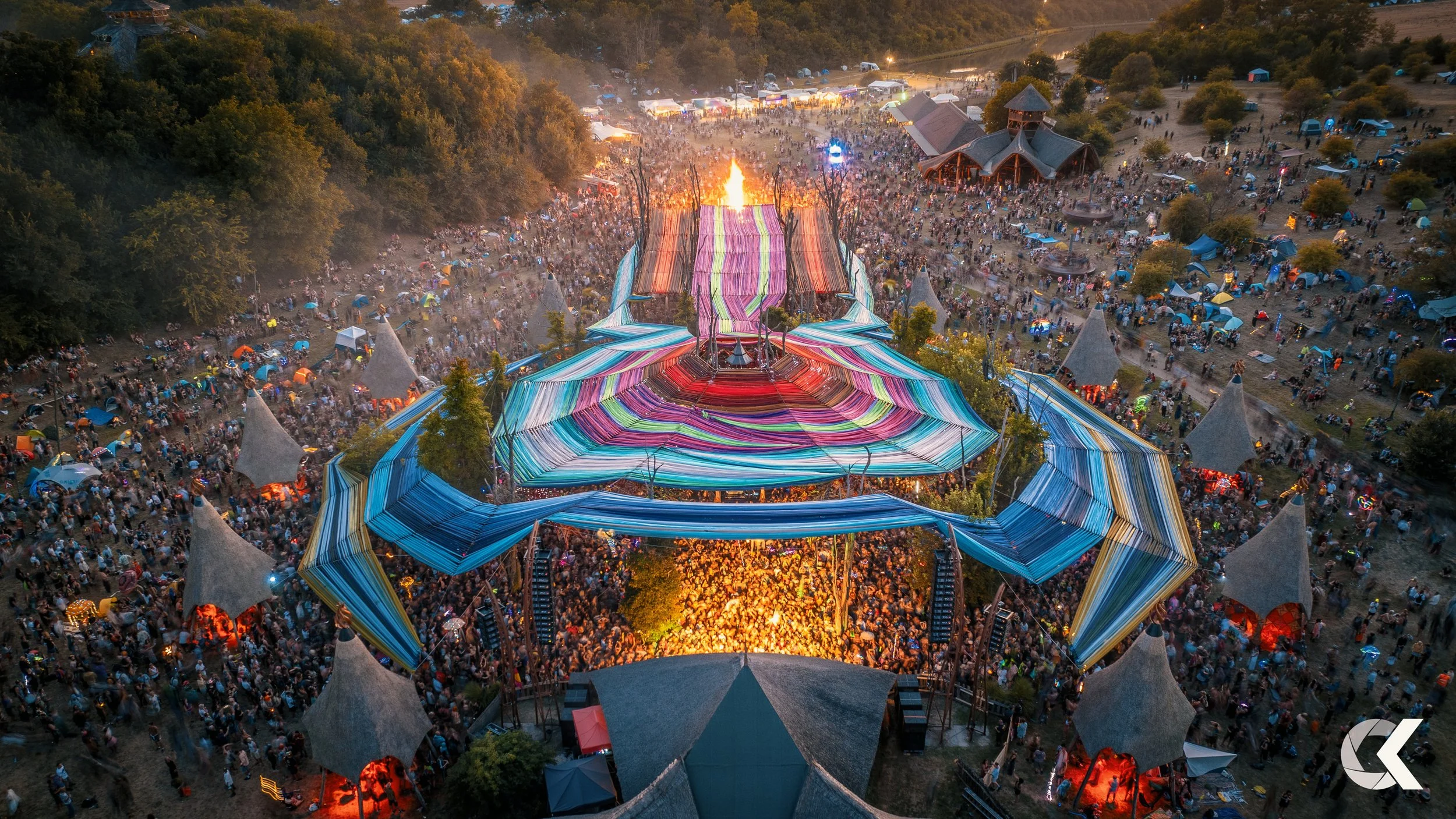 Aerial view of a large outdoor music festival with a colorful, spider-shaped stage, surrounded by a crowd of attendees and tents in a wooded area at sunset.