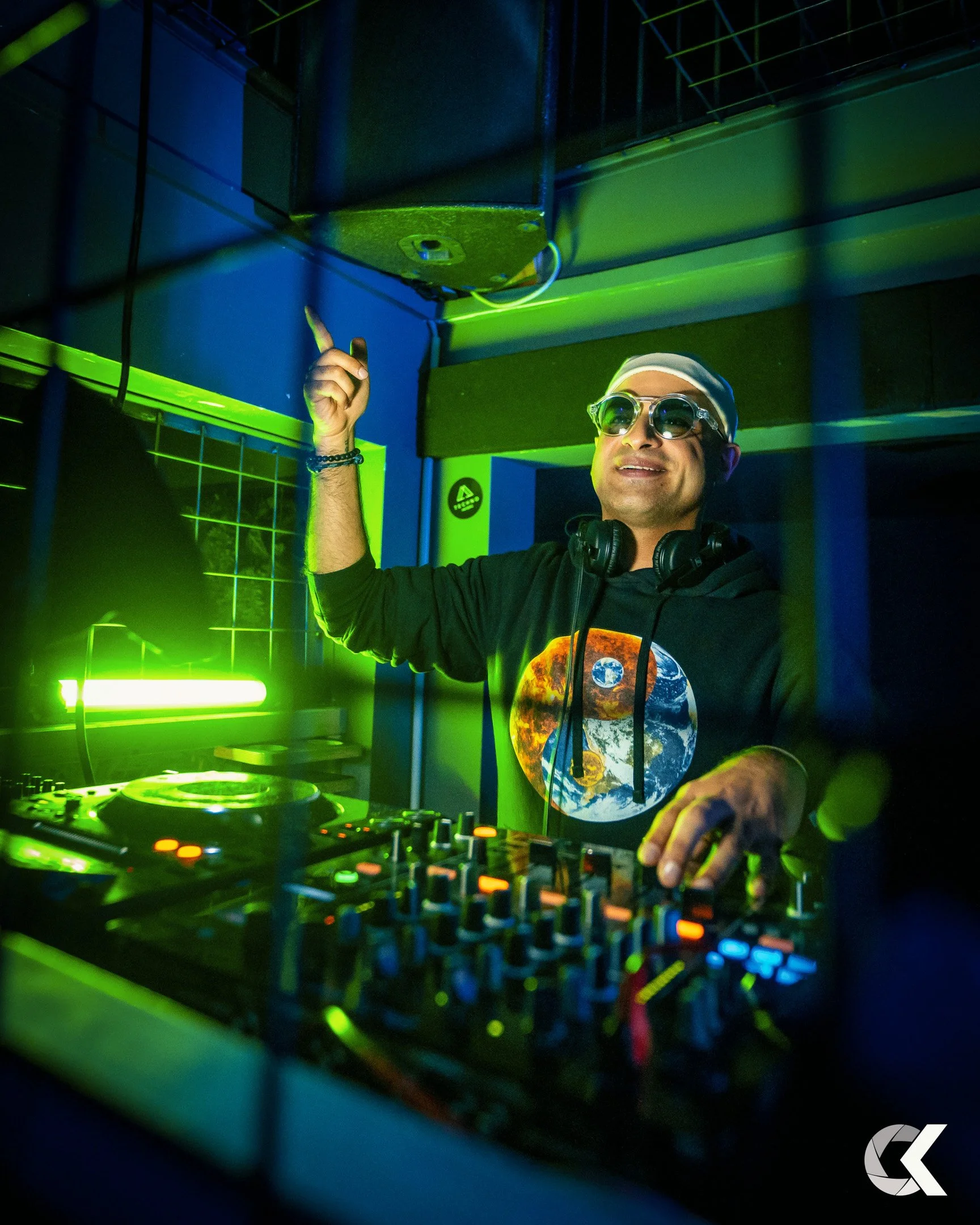 DJ wearing sunglasses and headphones, pointing upward while standing behind a DJ booth with colorful lights in a dark room.