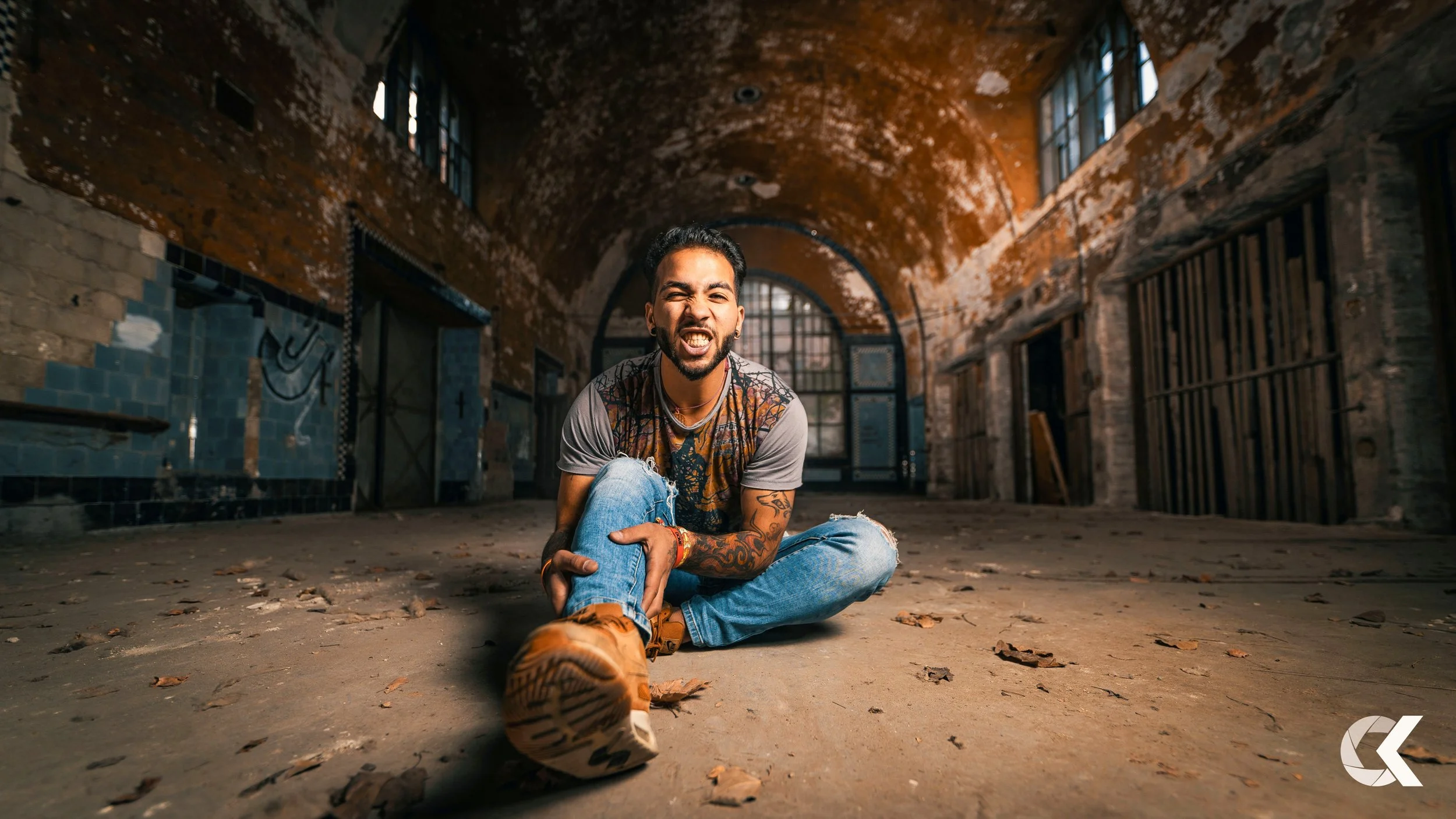A young man with tattoos and earrings sitting on the ground in an abandoned, industrial-style building, grimacing with one eye closed and mouth open, wearing a gray graphic t-shirt, ripped blue jeans, and tan shoes.