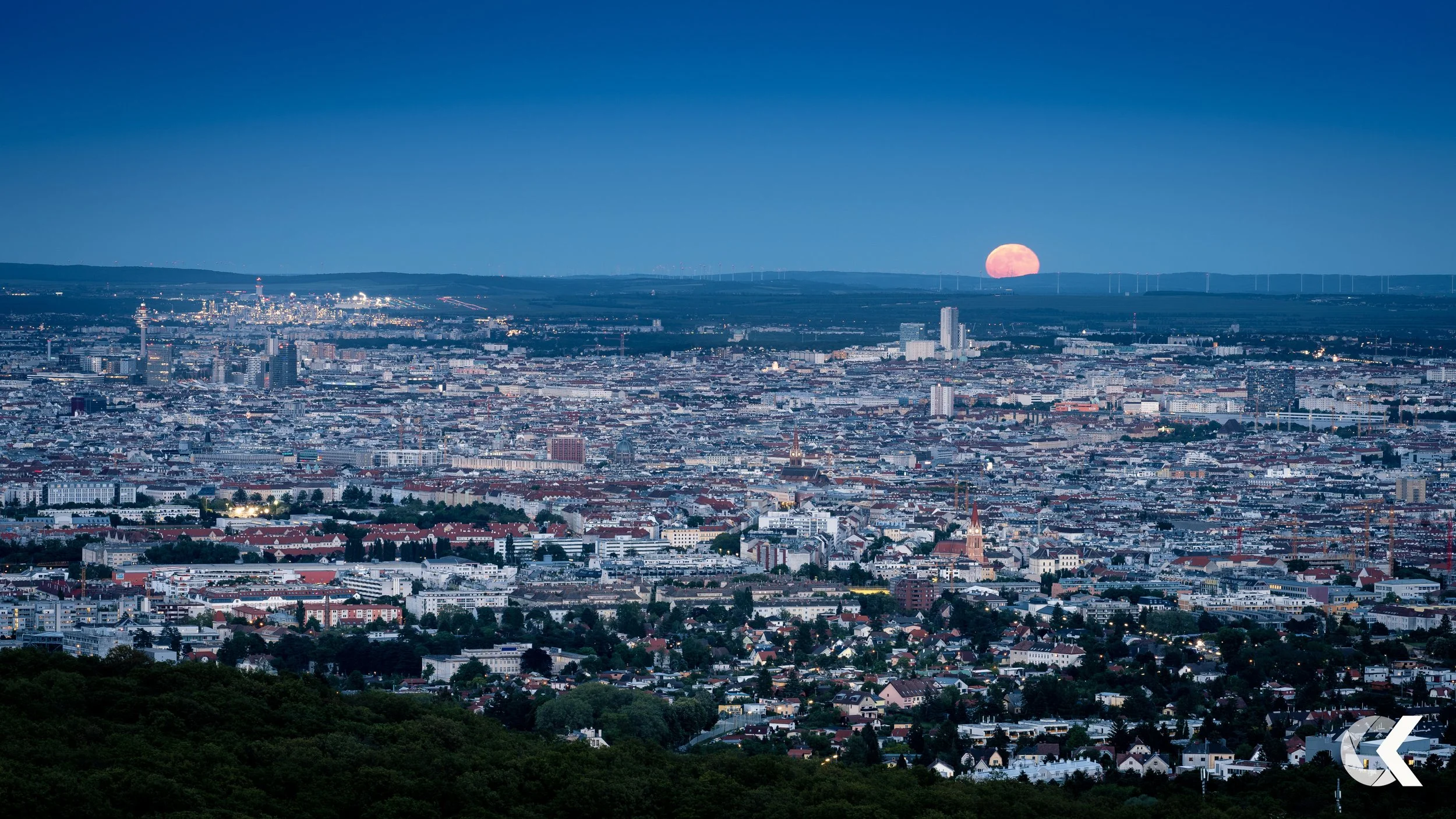 Cityscape at dusk with a large full moon rising above the horizon and a clear sky.