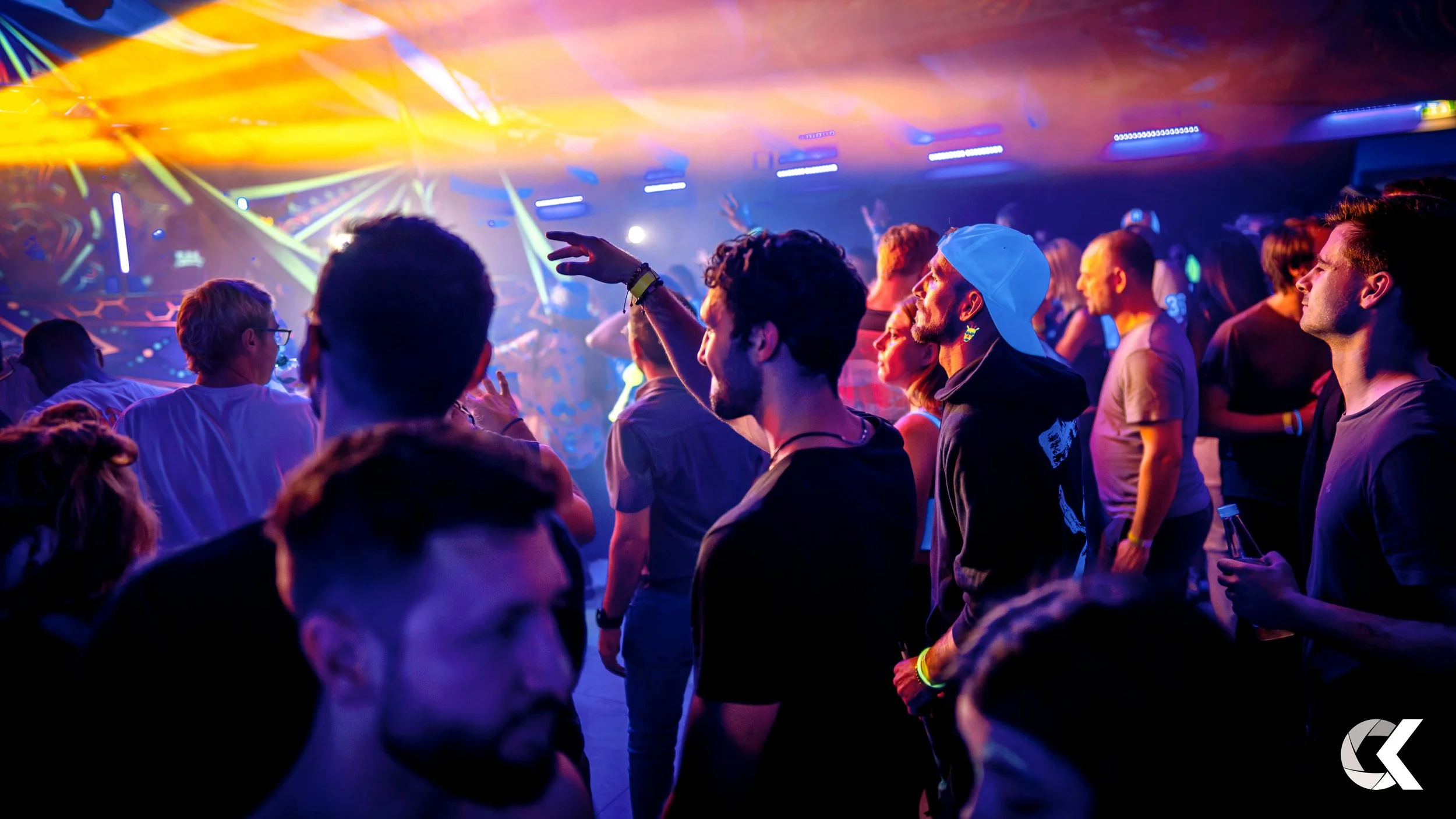 Crowd of people dancing and enjoying music at a concert or nightclub with colorful stage lights and lasers in the background.