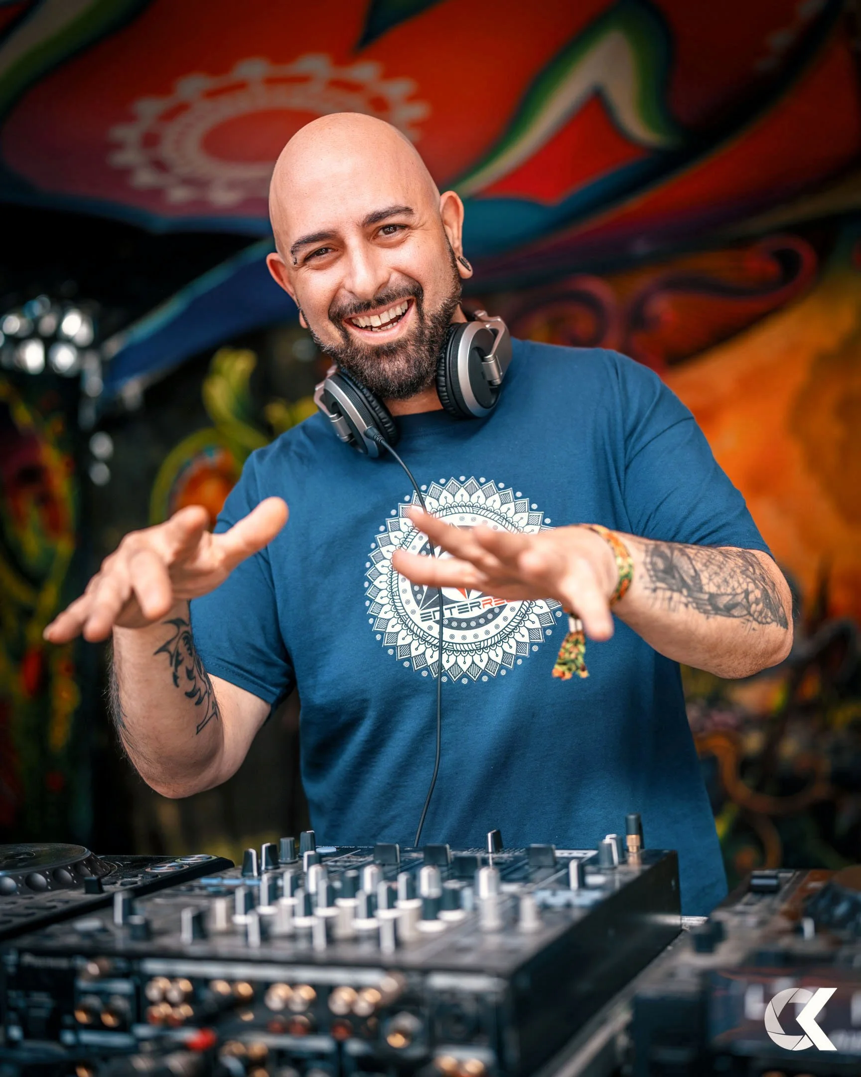 A smiling male DJ with headphones around his neck, standing behind a DJ mixer, gesturing with his hands, with colorful psychedelic art in the background.