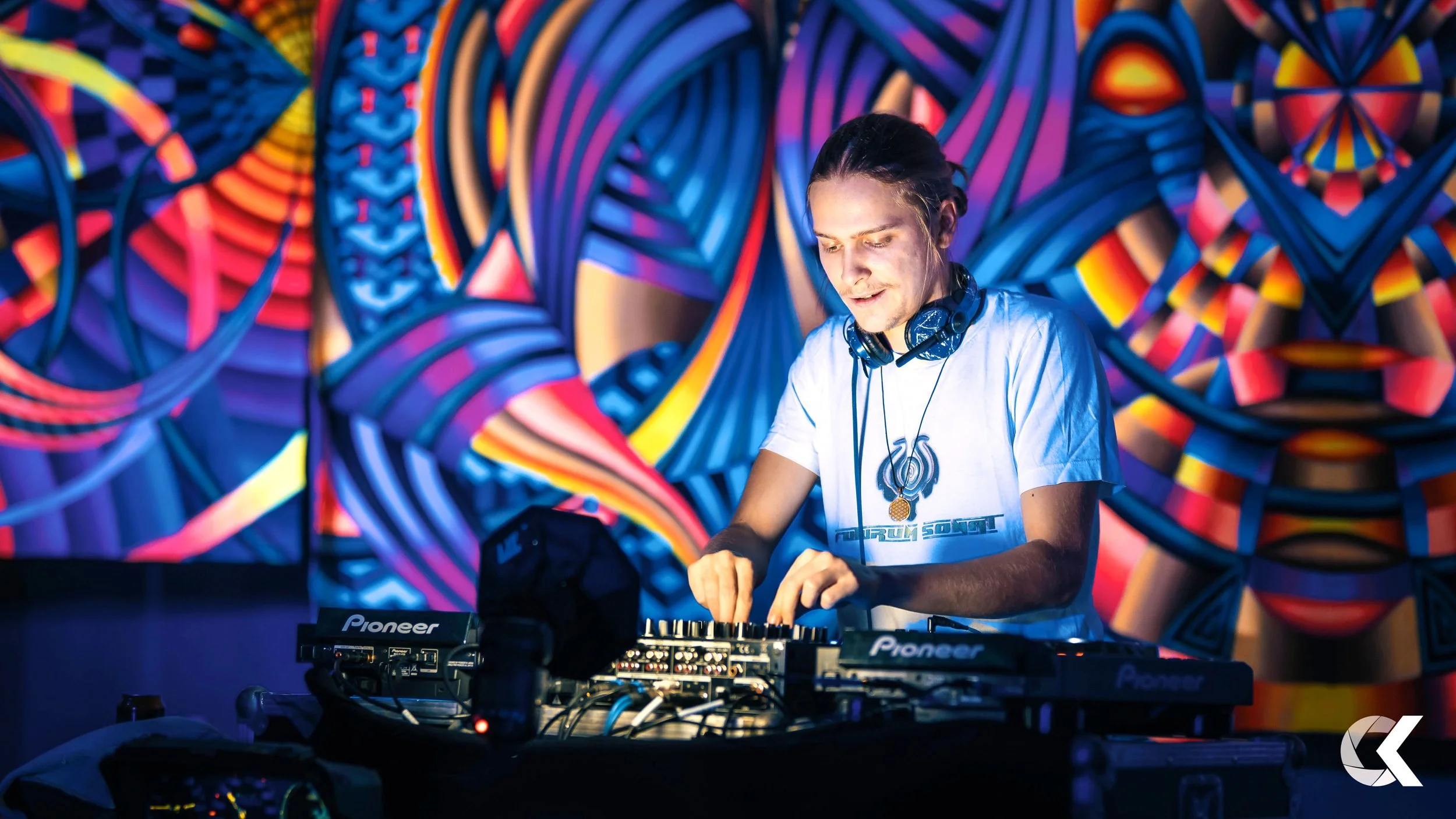 A young male DJ with headphones around his neck, wearing a white t-shirt, is mixing music on professional DJ equipment in front of a vibrant, colorful abstract mural.