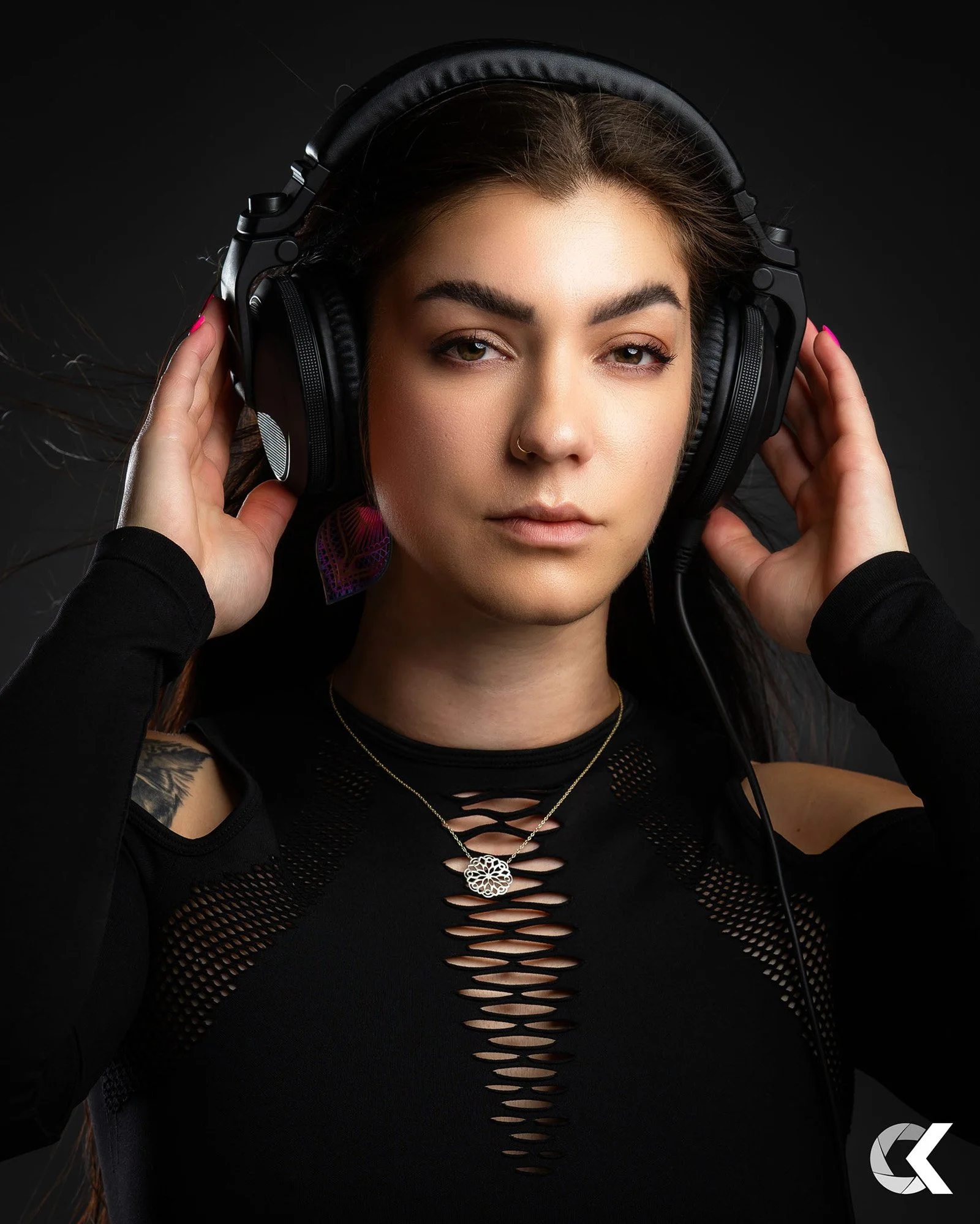 A woman with long brown hair, wearing a black top with cutouts and a necklace, holds large black headphones over her ears against a dark background.
