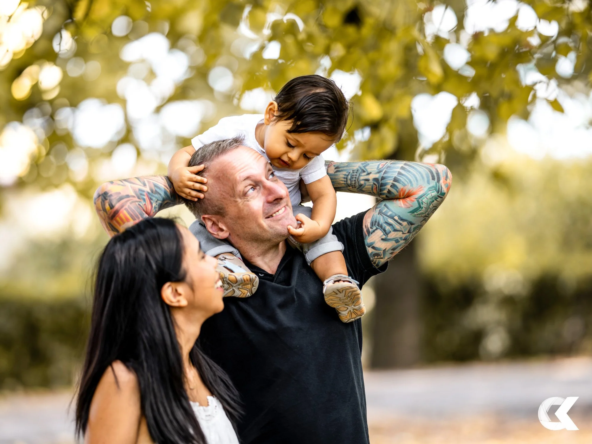 Family outdoors: father with tattoos carrying a young child on shoulders, mother looking at them, autumn trees in background.