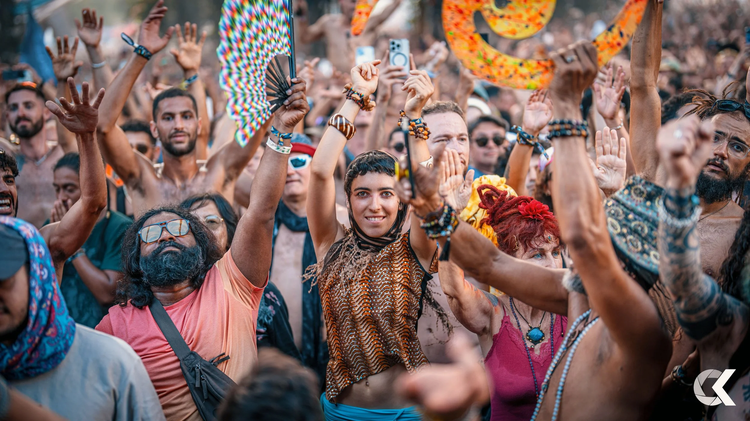 Crowd of people at a music festival or outdoor event, raising their hands, some holding accessories or phones, with a woman in the center smiling and wearing colorful bracelets.