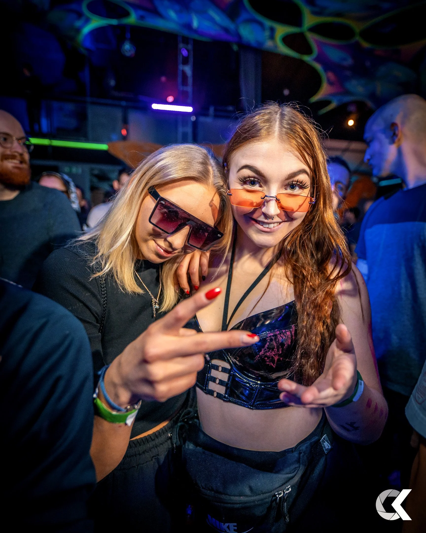 Two young women at a party or concert, both wearing sunglasses and smiling at the camera. They are surrounded by other people and colorful lighting.