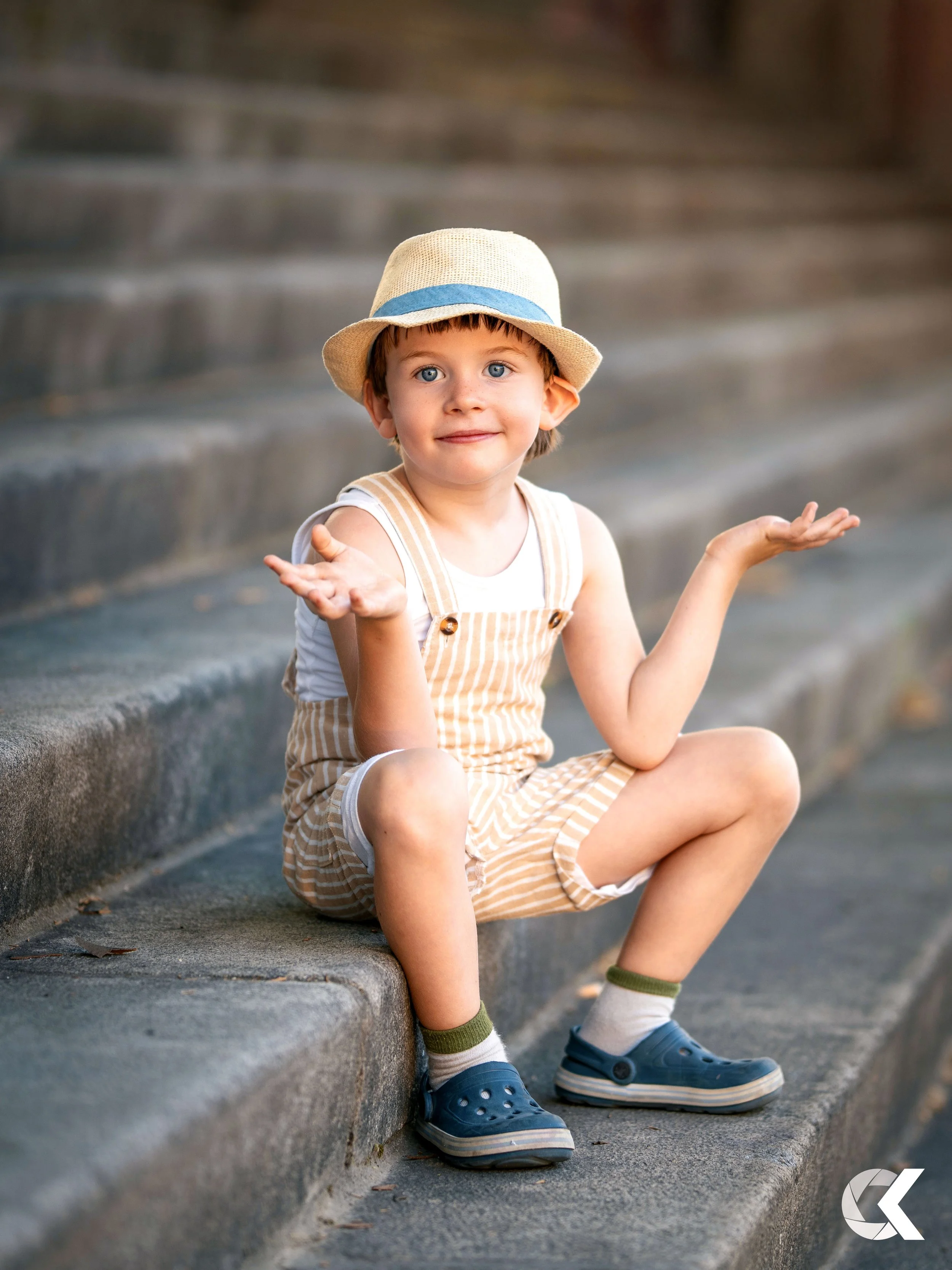 A young boy with blonde hair and blue eyes, wearing a beige hat with a blue band, a white sleeveless shirt, beige and white striped shorts, blue Crocs, and striped socks, sitting on concrete stairs with a puzzled or shrugging gesture.