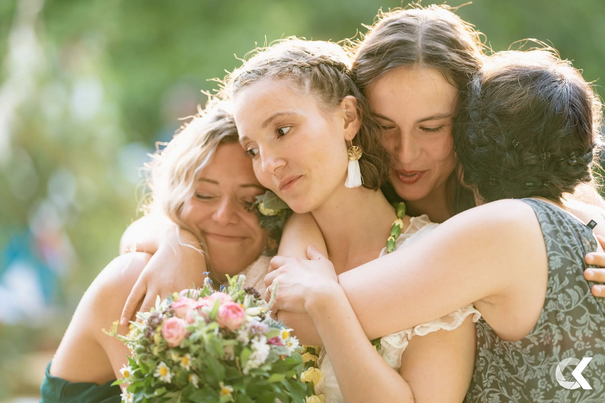 Four women hugging each other closely, with a woman in the center holding a bouquet of flowers, outdoors with green background.