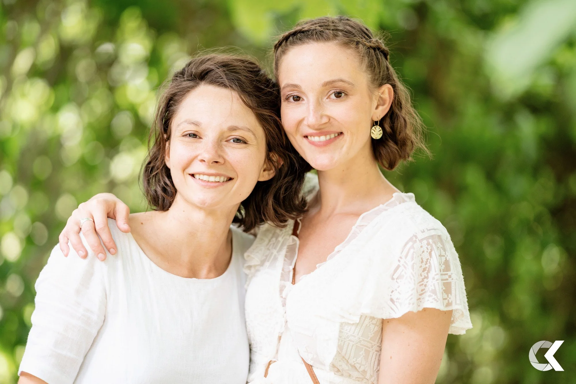 Two smiling women standing close together outdoors with a green, blurred background. One has short, dark wavy hair, and the other has medium-length, curly brown hair, wearing earrings. Both are wearing white tops.
