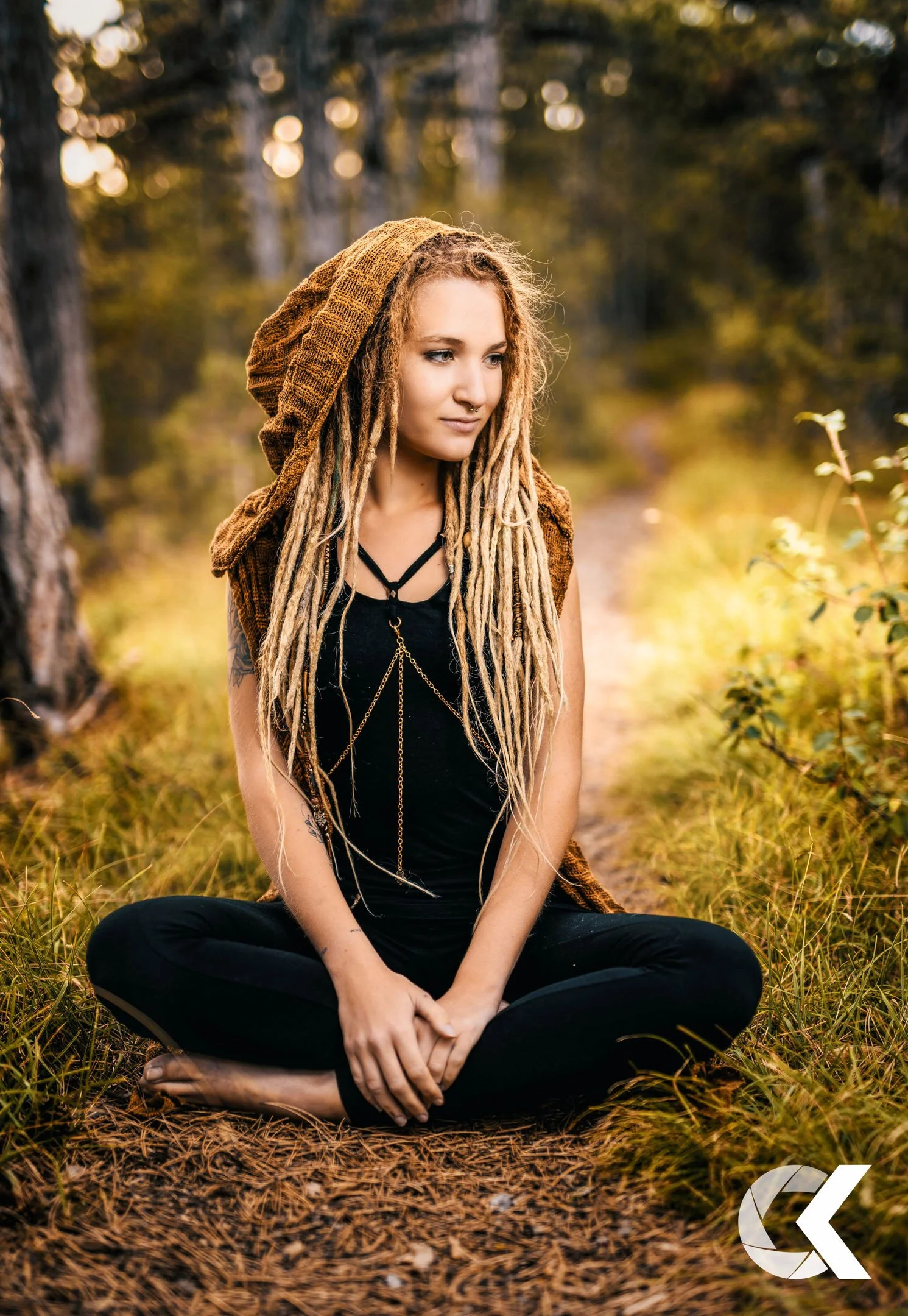 A young woman with blond dreadlocks sitting cross-legged on a forest trail during sunset, wearing a black top, brown vest, and layered necklaces.